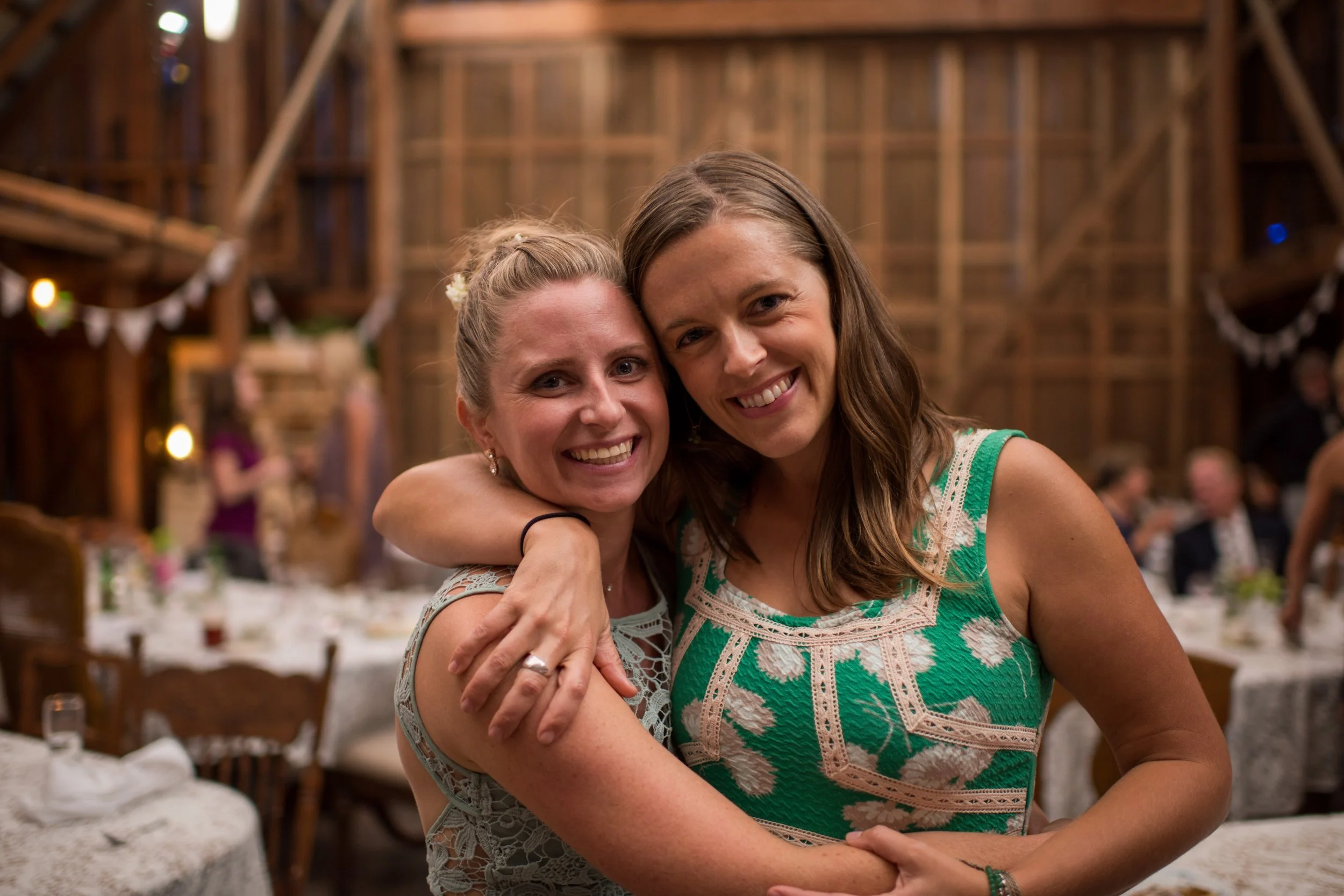 Two women smiling and hugging each other at a social gathering inside a wooden barn with string lights and decorated tables.