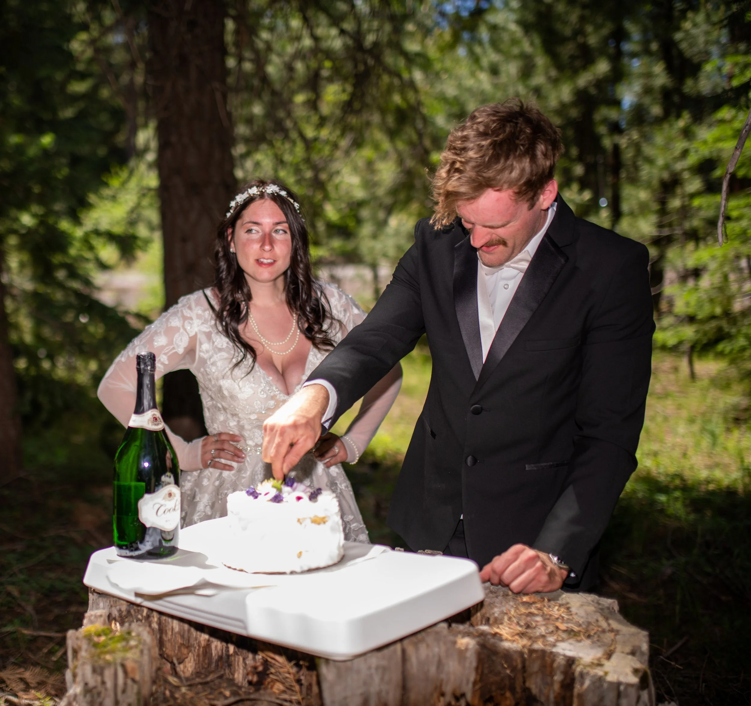 A wedding cake on a white table, outdoors in a wooded area, with a man in a tuxedo cutting the cake and a woman in a white dress with a floral headband watching.