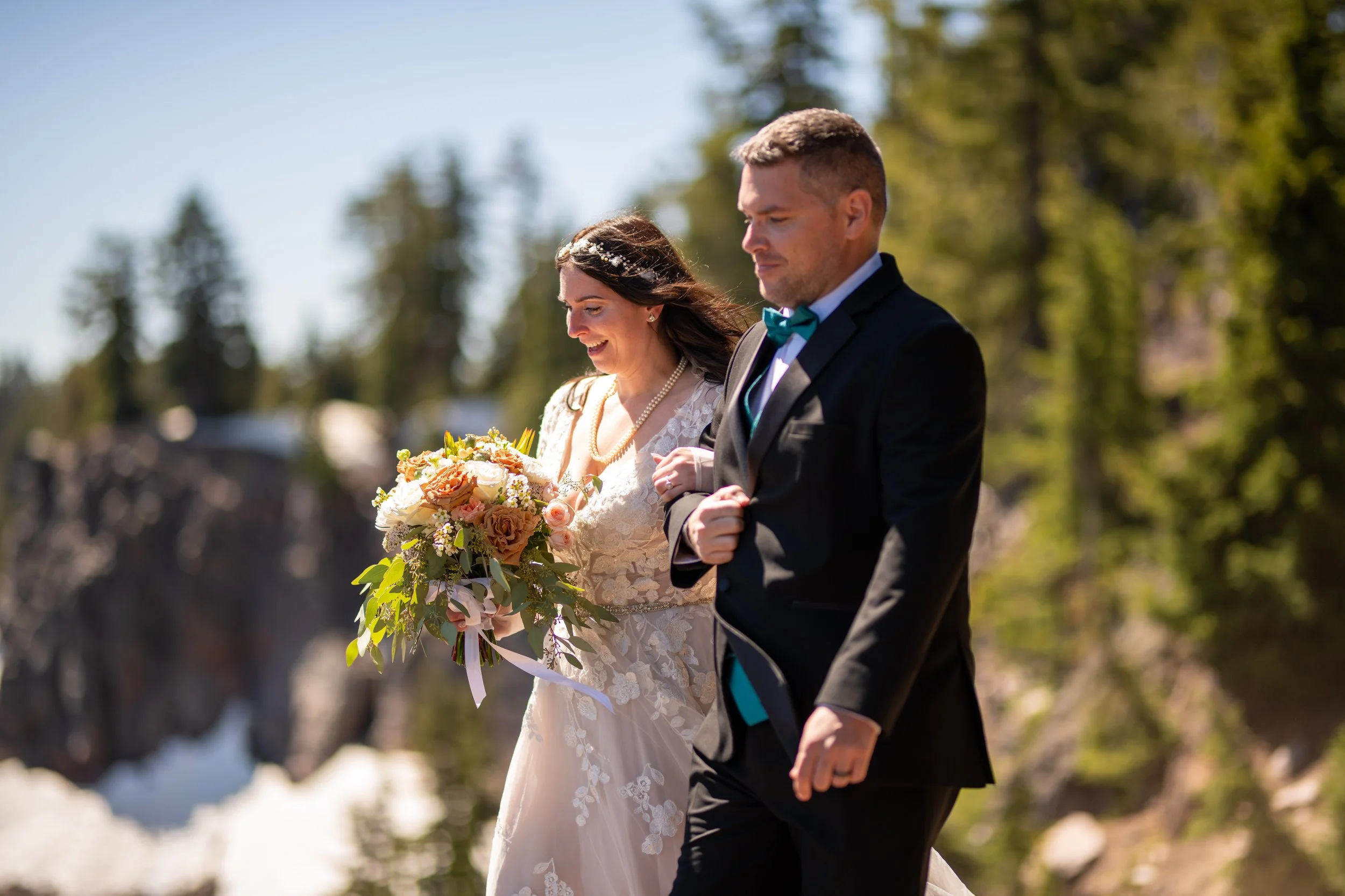A bride and groom walking outdoors during a wedding ceremony, with the bride holding a bouquet of flowers and wearing a lace wedding dress, while the groom is dressed in a black suit with a teal bow tie and holding her arm.