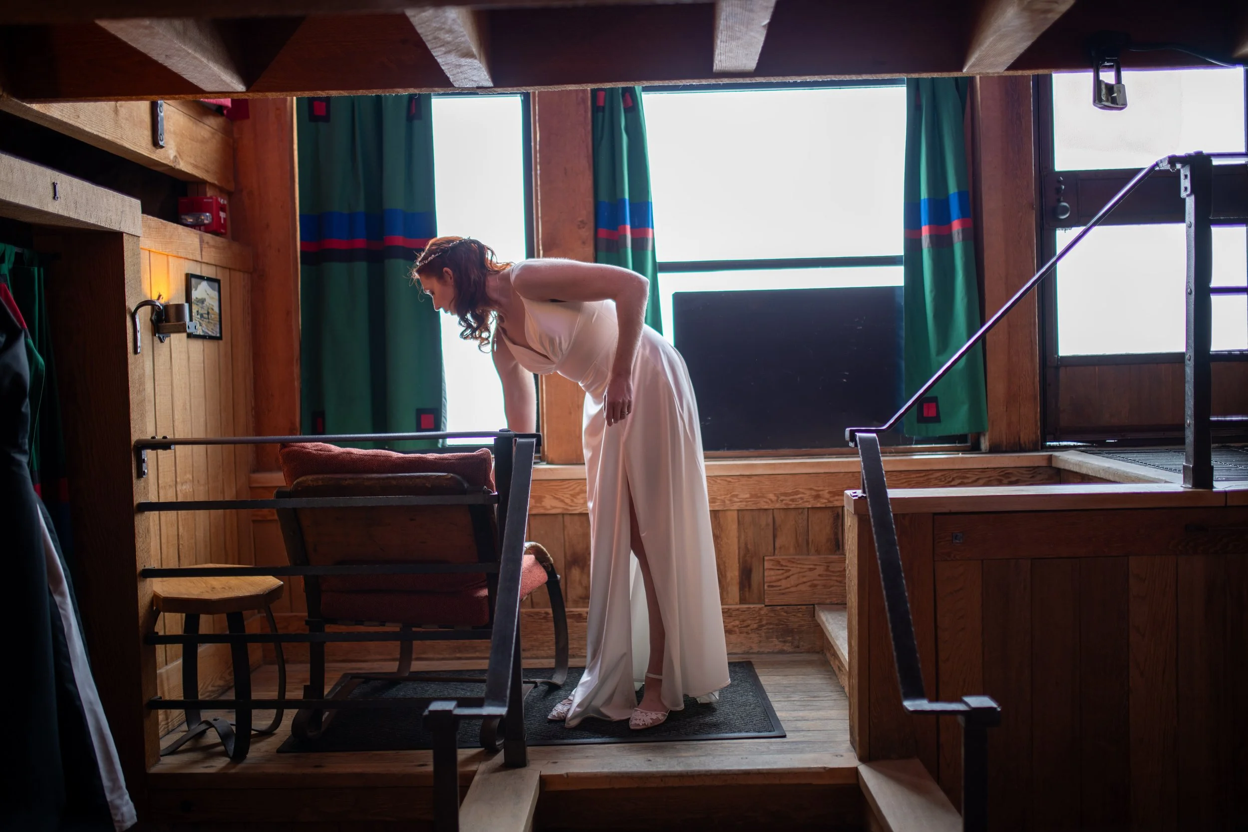 A woman in a white dress standing at the top of a staircase in a room with wooden walls, green curtains, and large windows letting in sunlight.