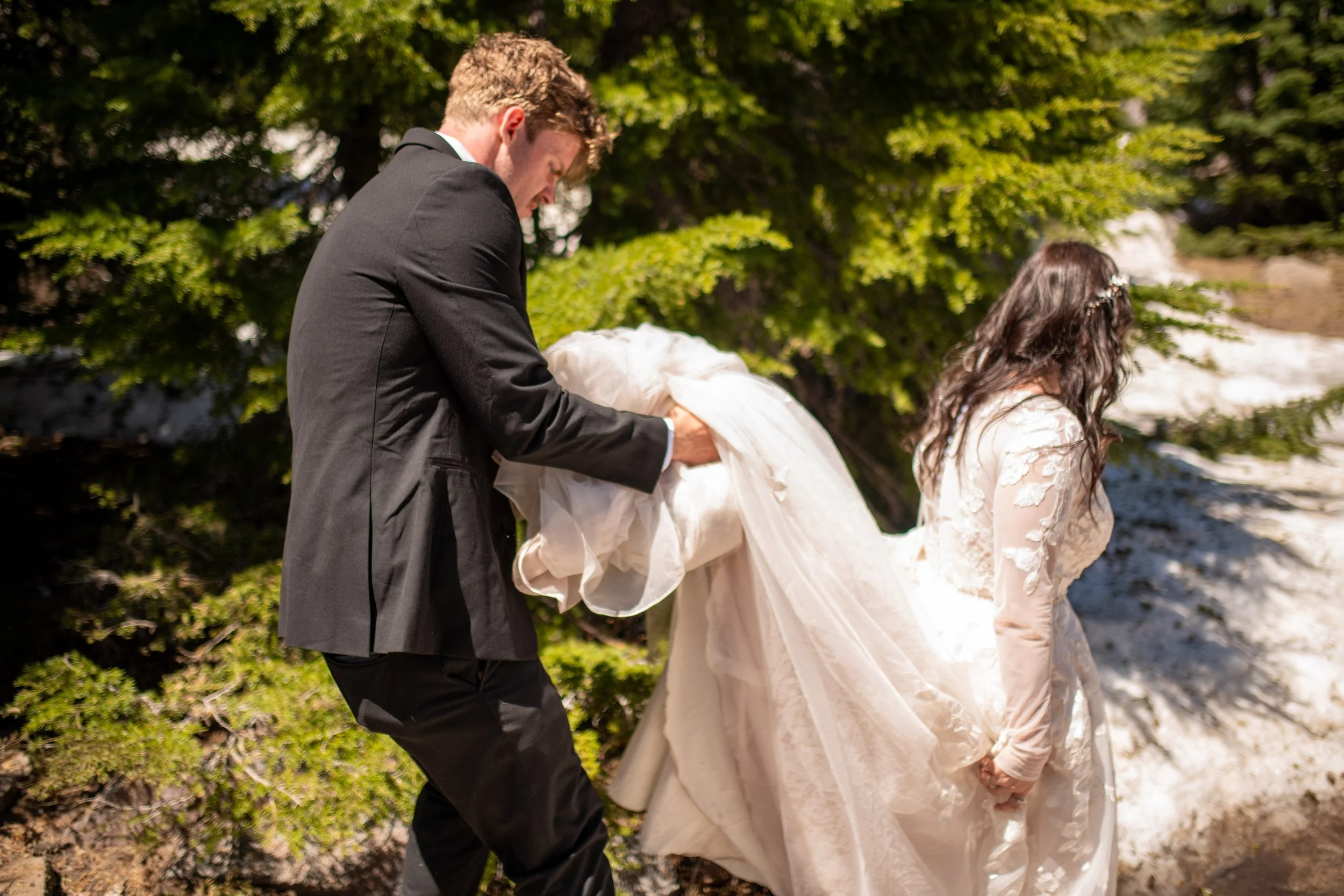 A man in a black suit and a woman in a white lace dress are outdoors near water, holding a wedding dress together, with green trees in the background.