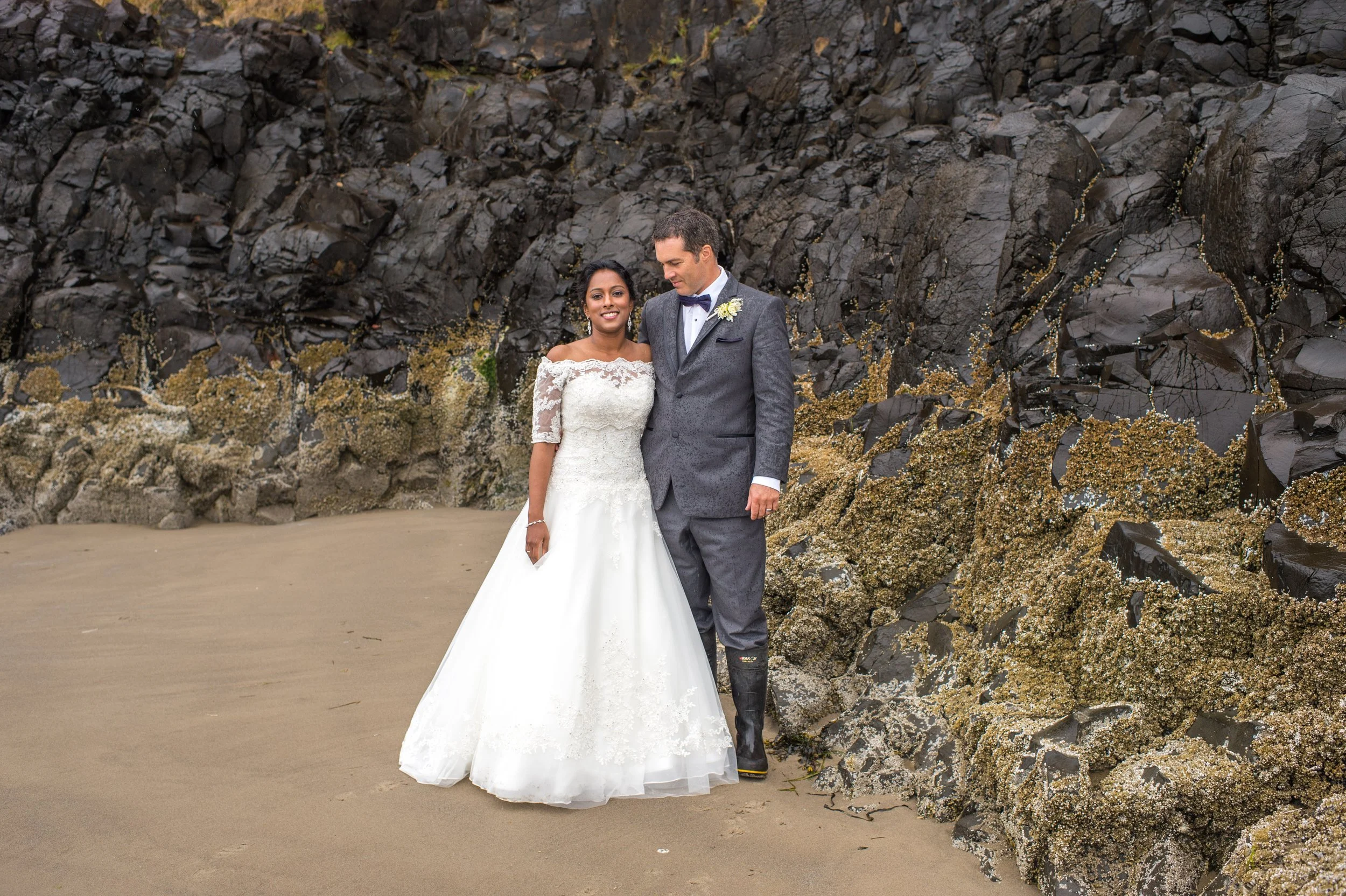 A bride and groom standing on a sandy beach with a large rocky cliff in the background. The bride is wearing a white wedding gown with lace details, and the groom is dressed in a gray suit with a bow tie. They are smiling and looking at each other.