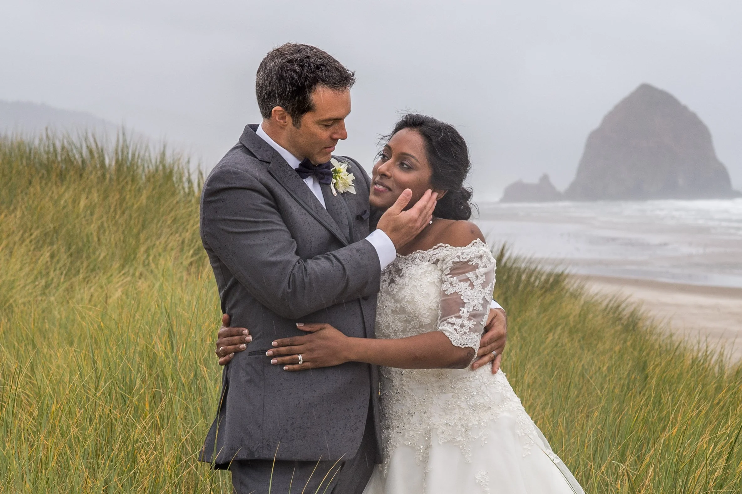 A newlywed couple embracing on a beach with grassy dunes in the foreground and large rock formations in the background. The groom is wearing a dark gray suit with a bow tie, and the bride is wearing a white lace wedding dress. It is raining lightly.