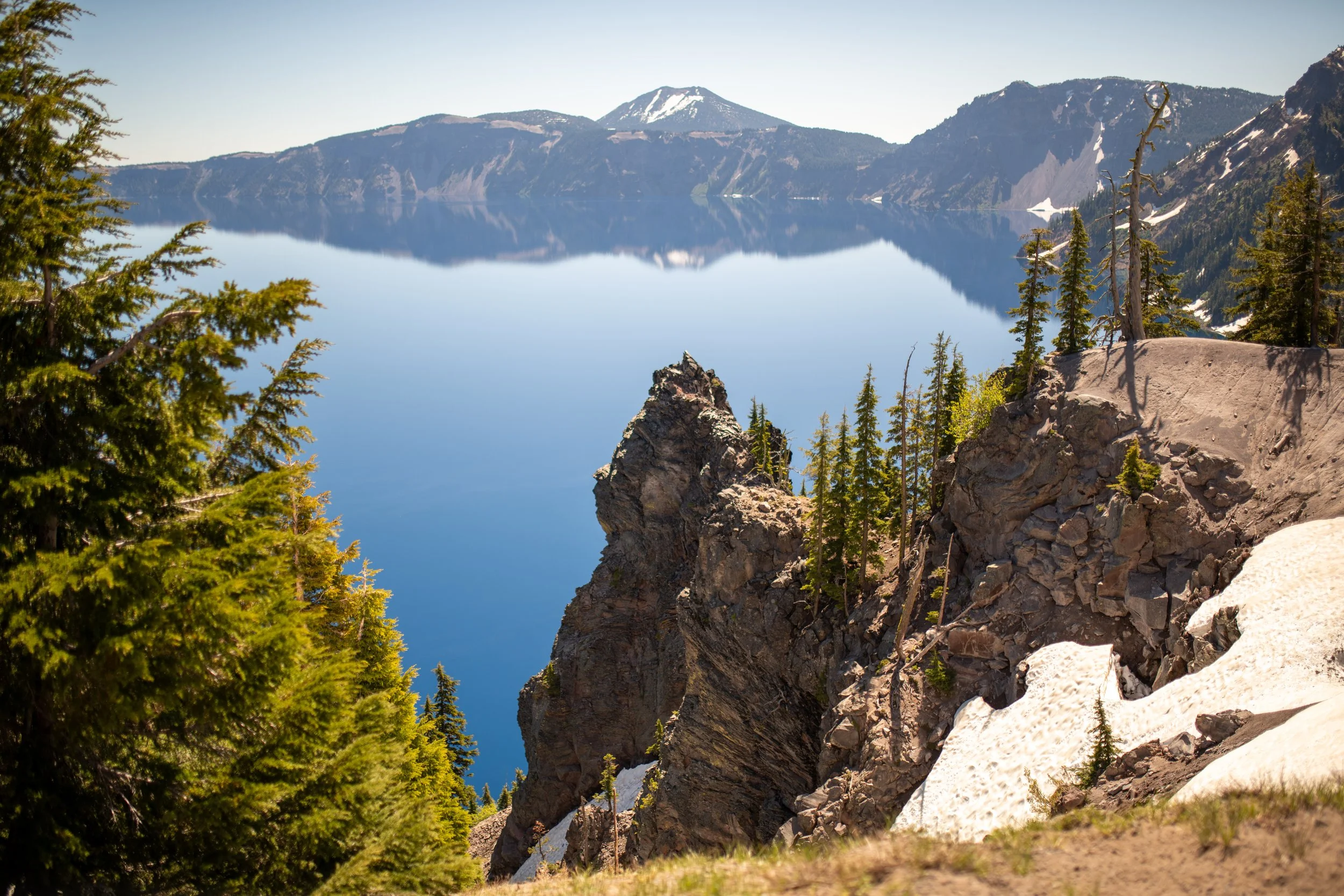 A calm mountain lake reflecting snow-capped peaks and a clear blue sky, with trees and rocky cliffs in the foreground.