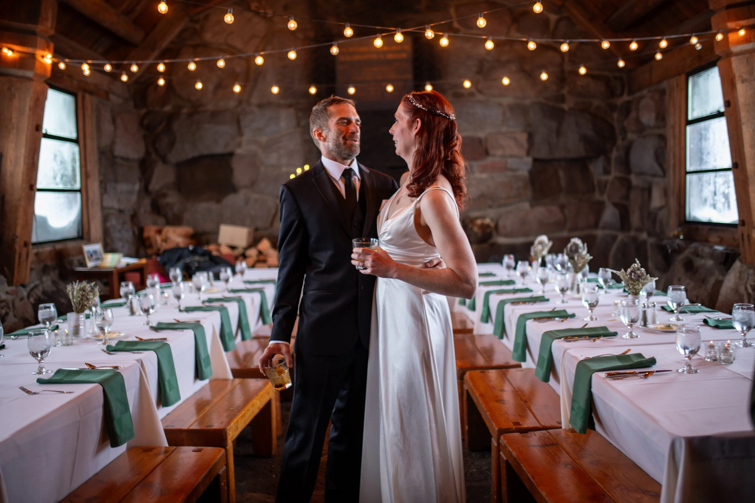 A bride and groom dancing at their wedding reception inside a rustic cabin with string lights overhead and tables set with white tablecloths and green napkins.