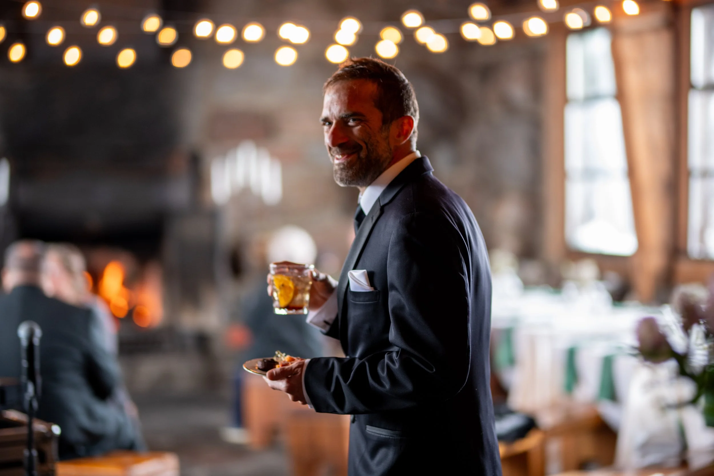 A man in a tuxedo holding a drink with a lemon wedge, smiling at a social event in a warmly lit venue.