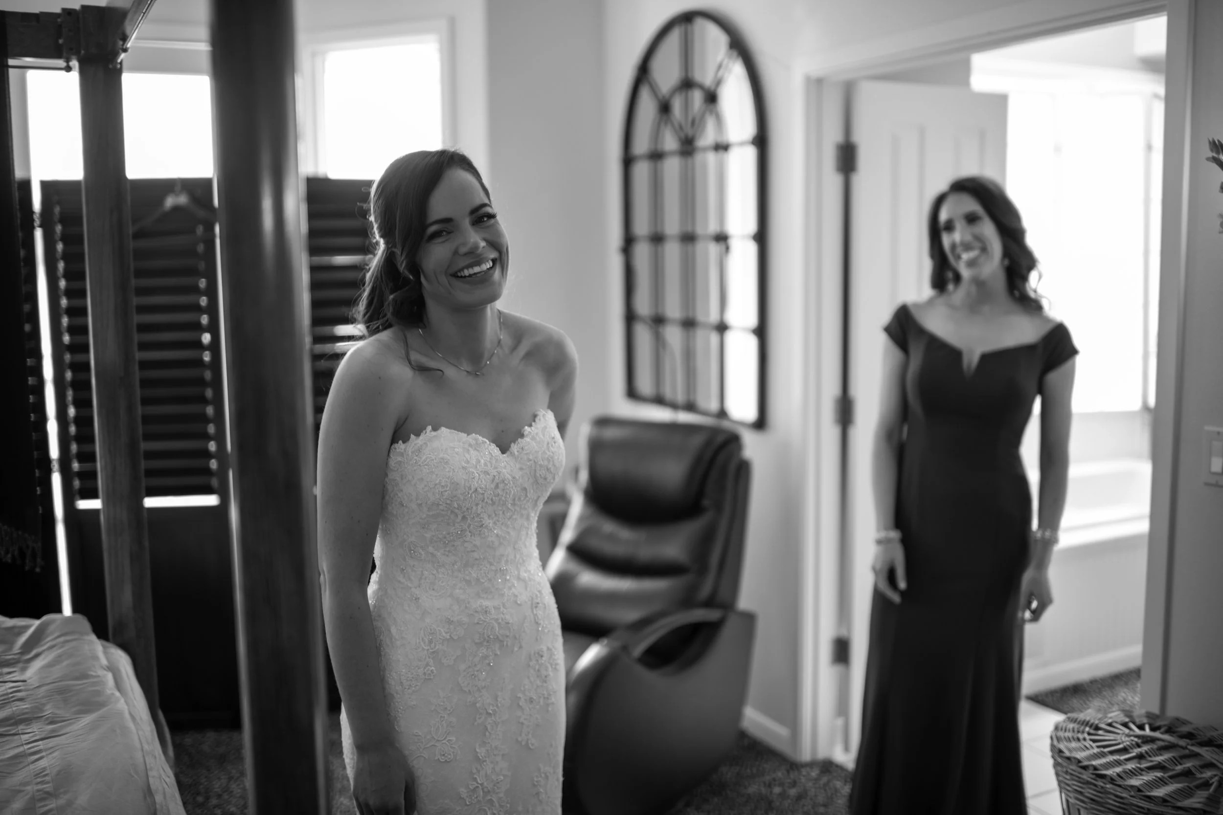A woman in a lace wedding dress smiling inside a room, with another woman in a dark dress in the background, near a window and mirror.