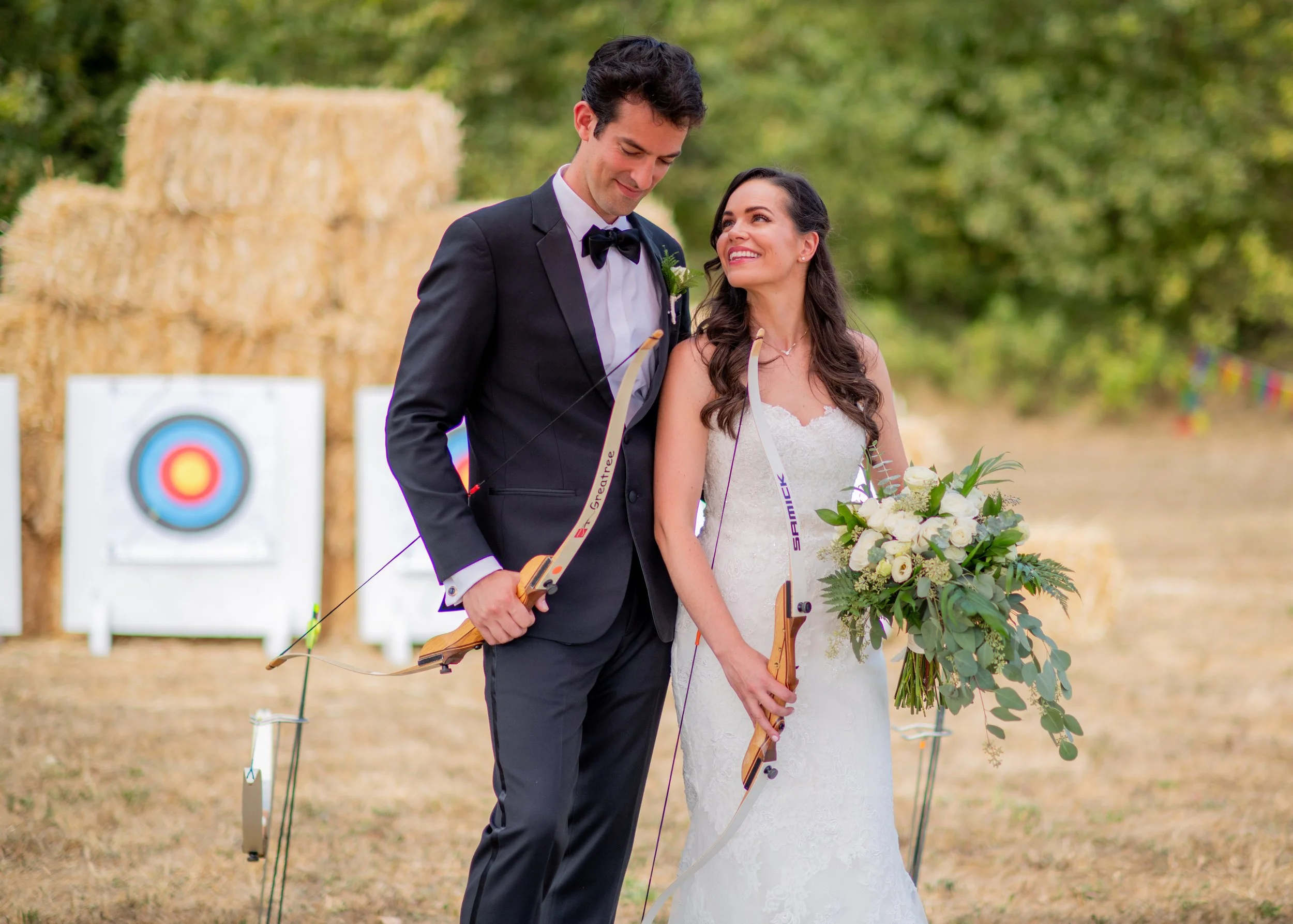 A bride and groom stand outdoors, smiling and looking at each other, holding bows and a bouquet of flowers, during a wedding ceremony with archery-themed decoration.