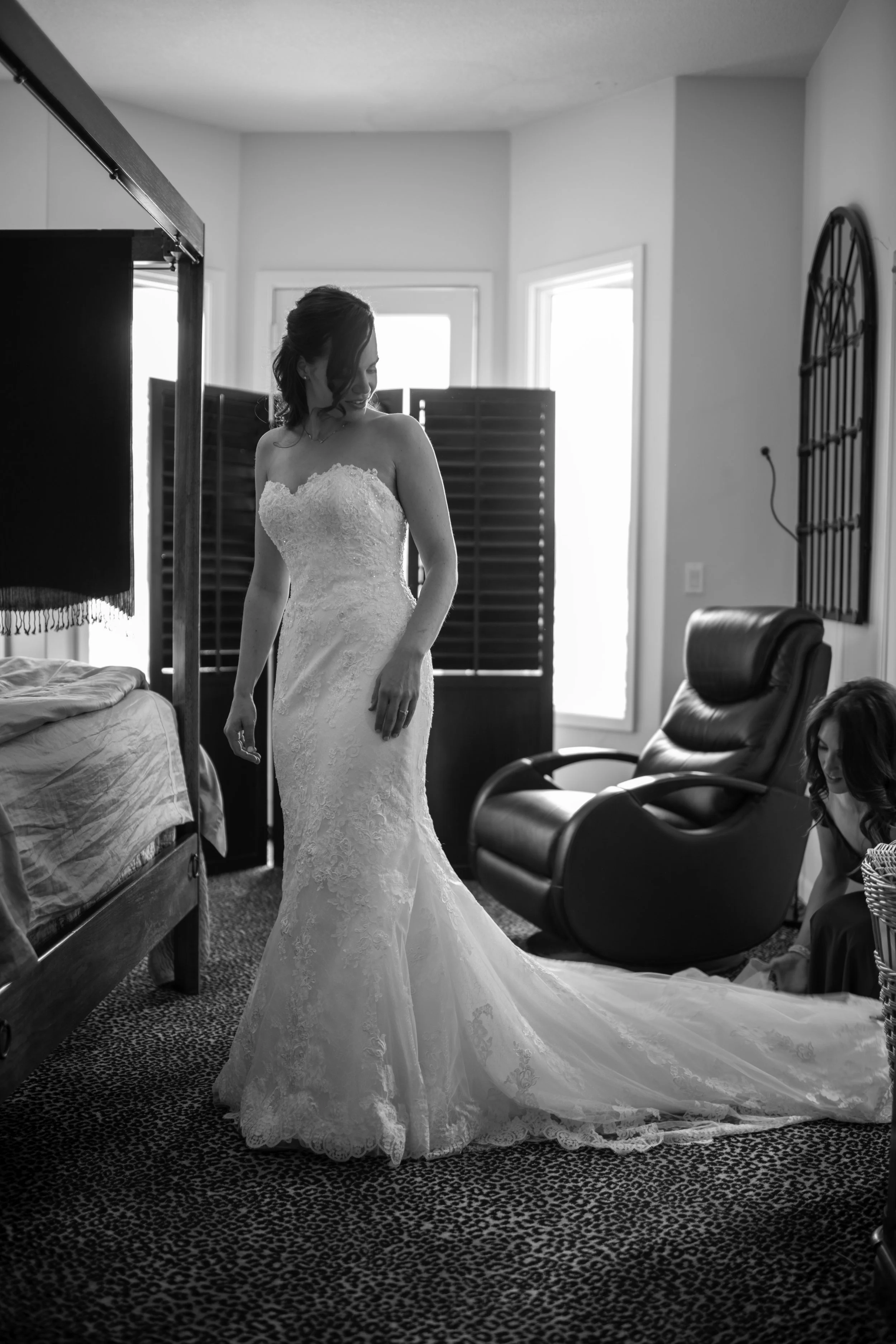 A bride in a strapless lace wedding gown standing beside a bed in a room, with a black leather chair and window in the background.