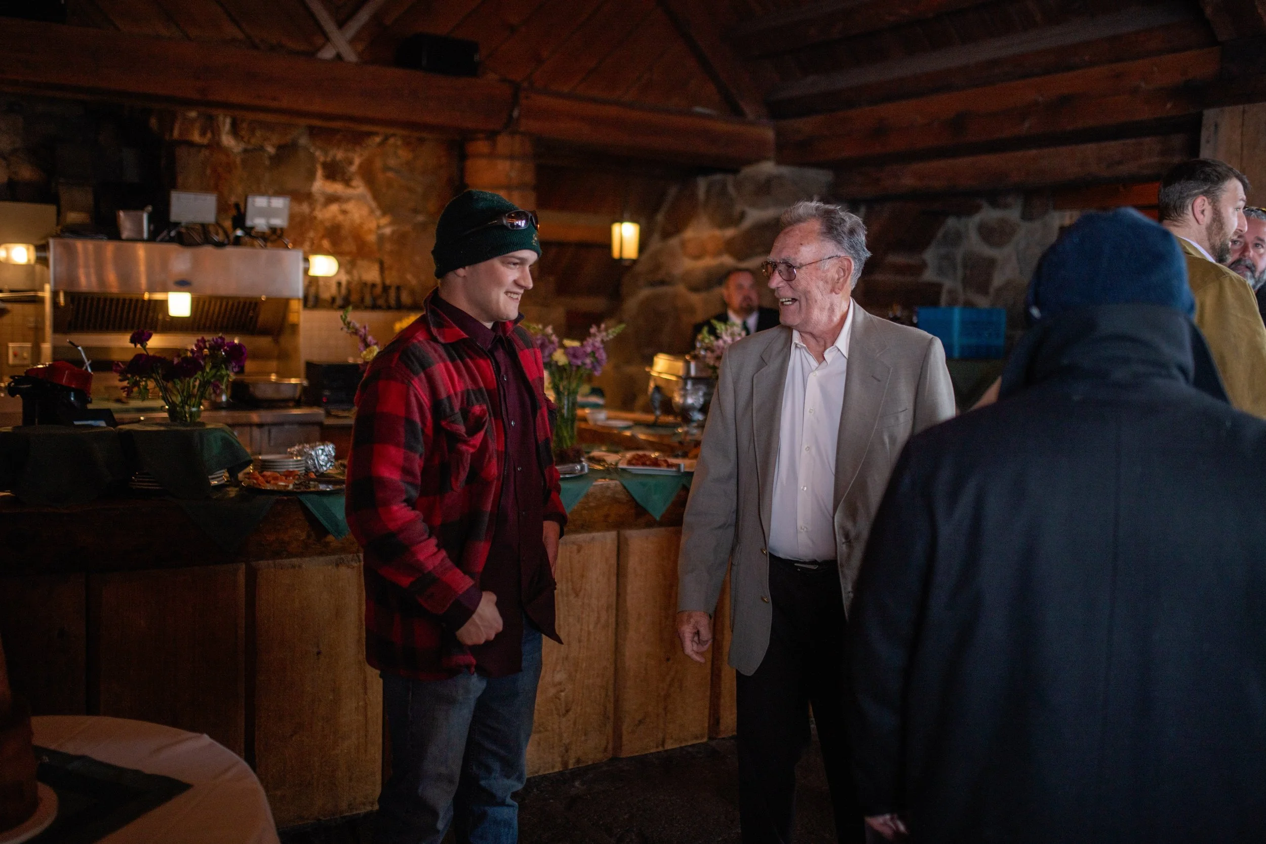 Group of people talking in a rustic kitchen or dining area with wooden walls and ceiling. There are flowers on the counter. The focus is on a young man in a black beanie and red plaid shirt and an elderly man in glasses and a beige suit jacket.