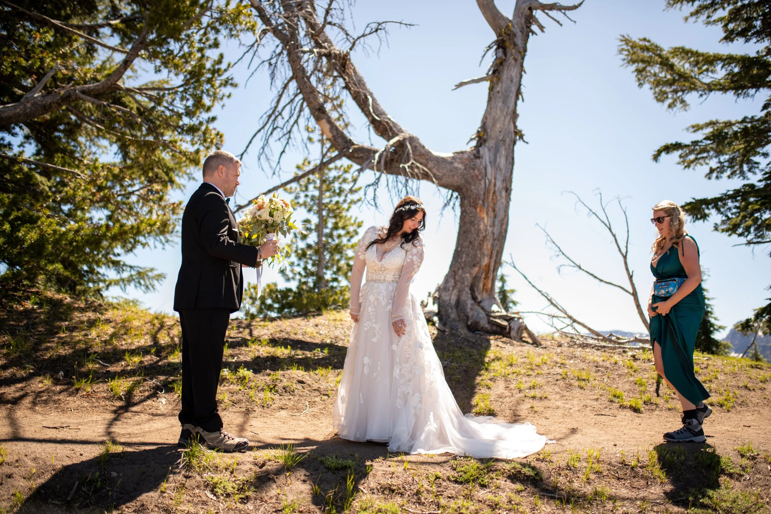 A bride in a white wedding dress and a bridegroom in a black suit standing outdoors under a large tree at a wedding ceremony, with an officiant or witness on the right, in a sunny, natural setting with other trees in the background.