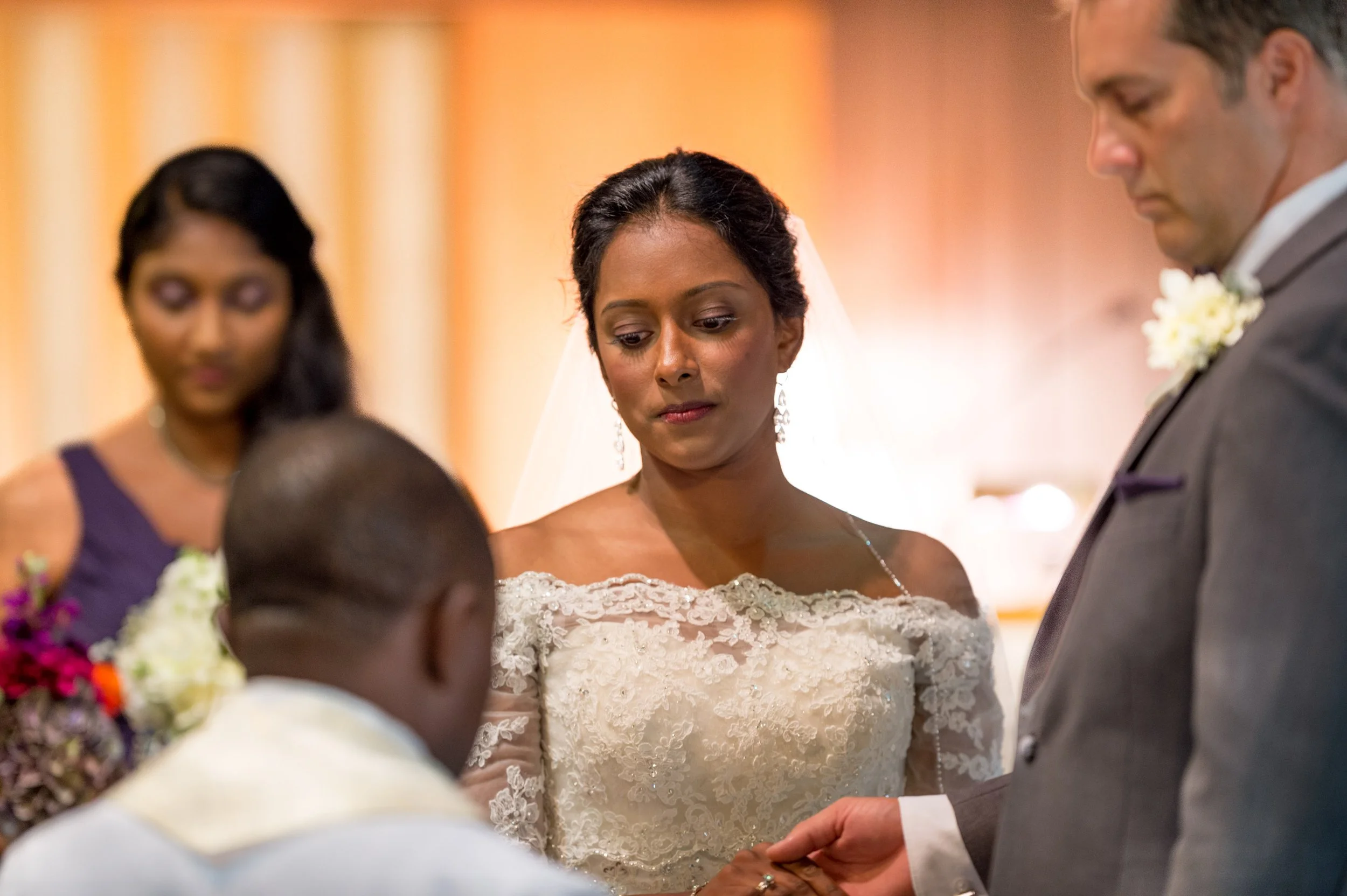 A bride and groom exchange wedding vows during a ceremony, with a woman and others in the background.