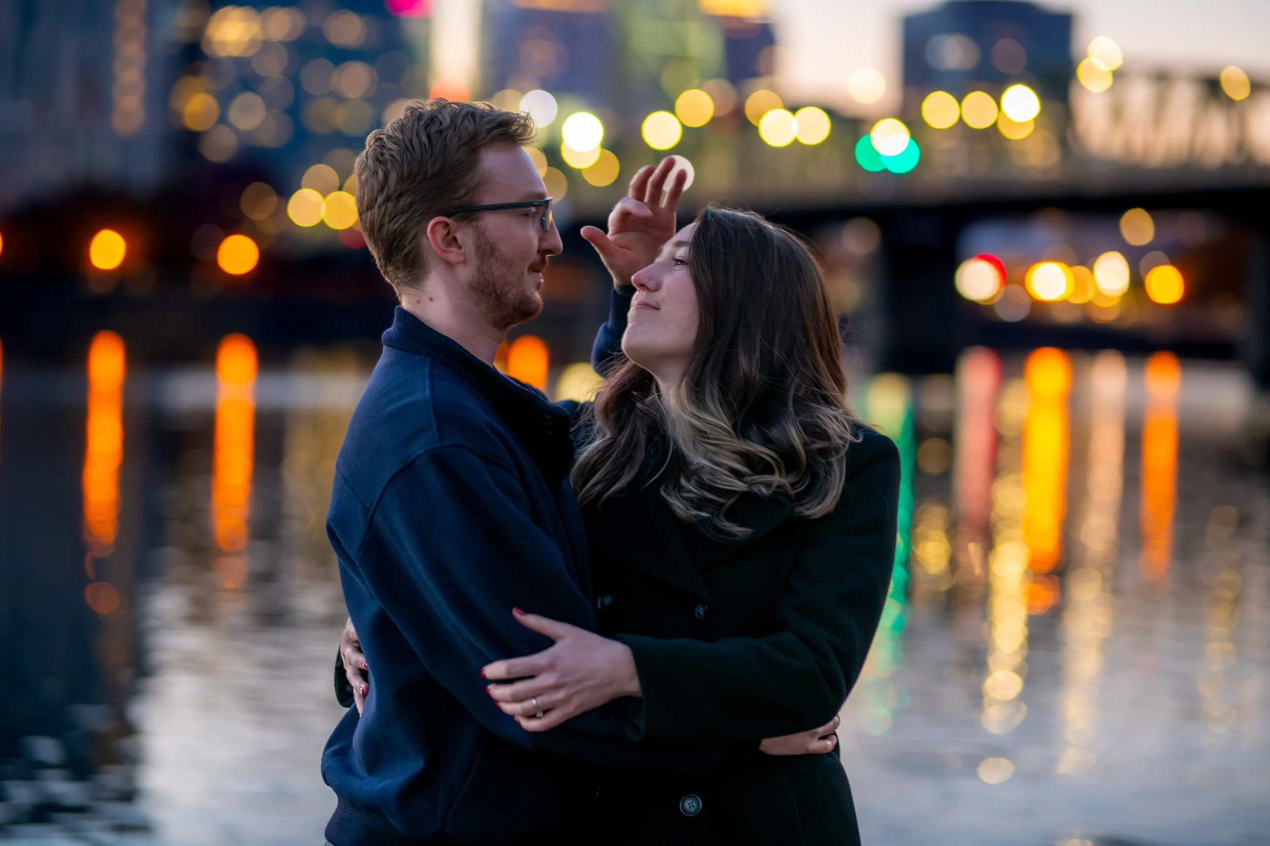 A couple sharing a close, intimate moment by a waterfront at dusk, with city lights reflecting on the water in the background.