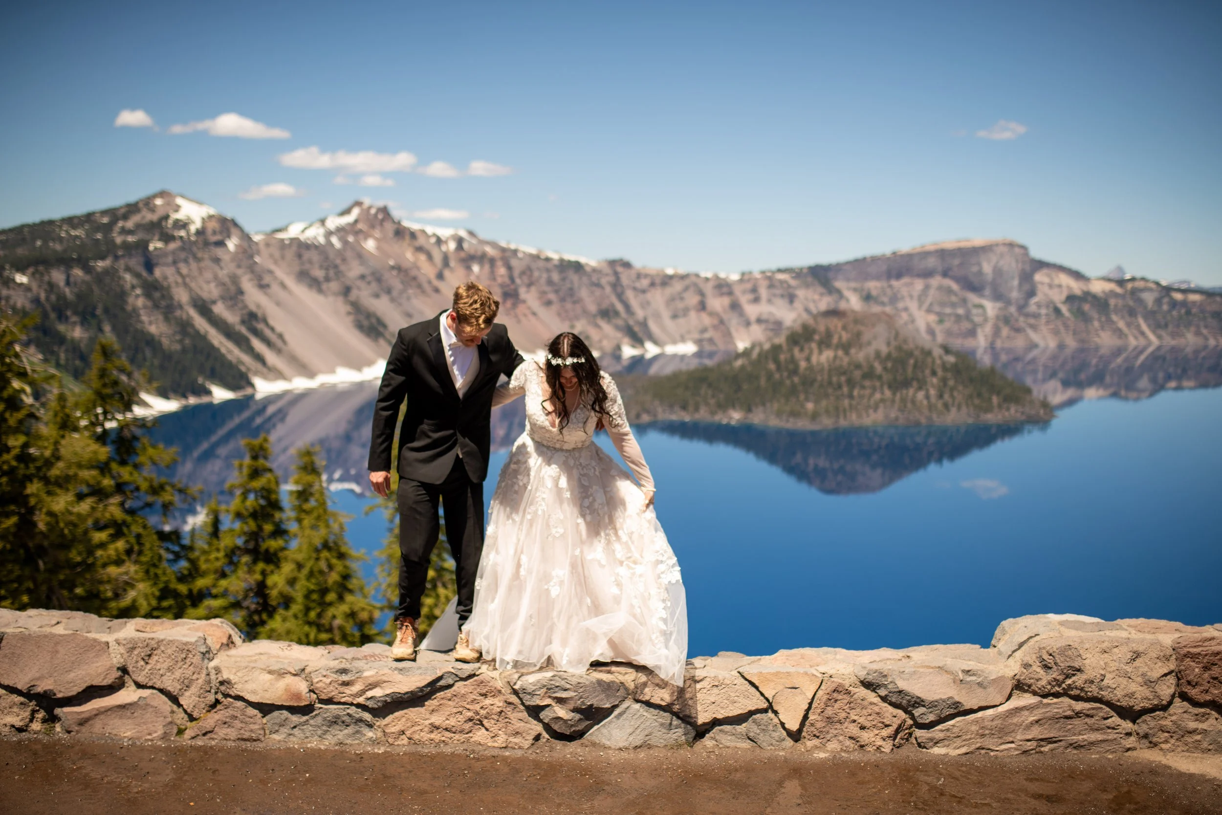 A bride and groom walking together near a lake with mountains in the background, during a sunny day.