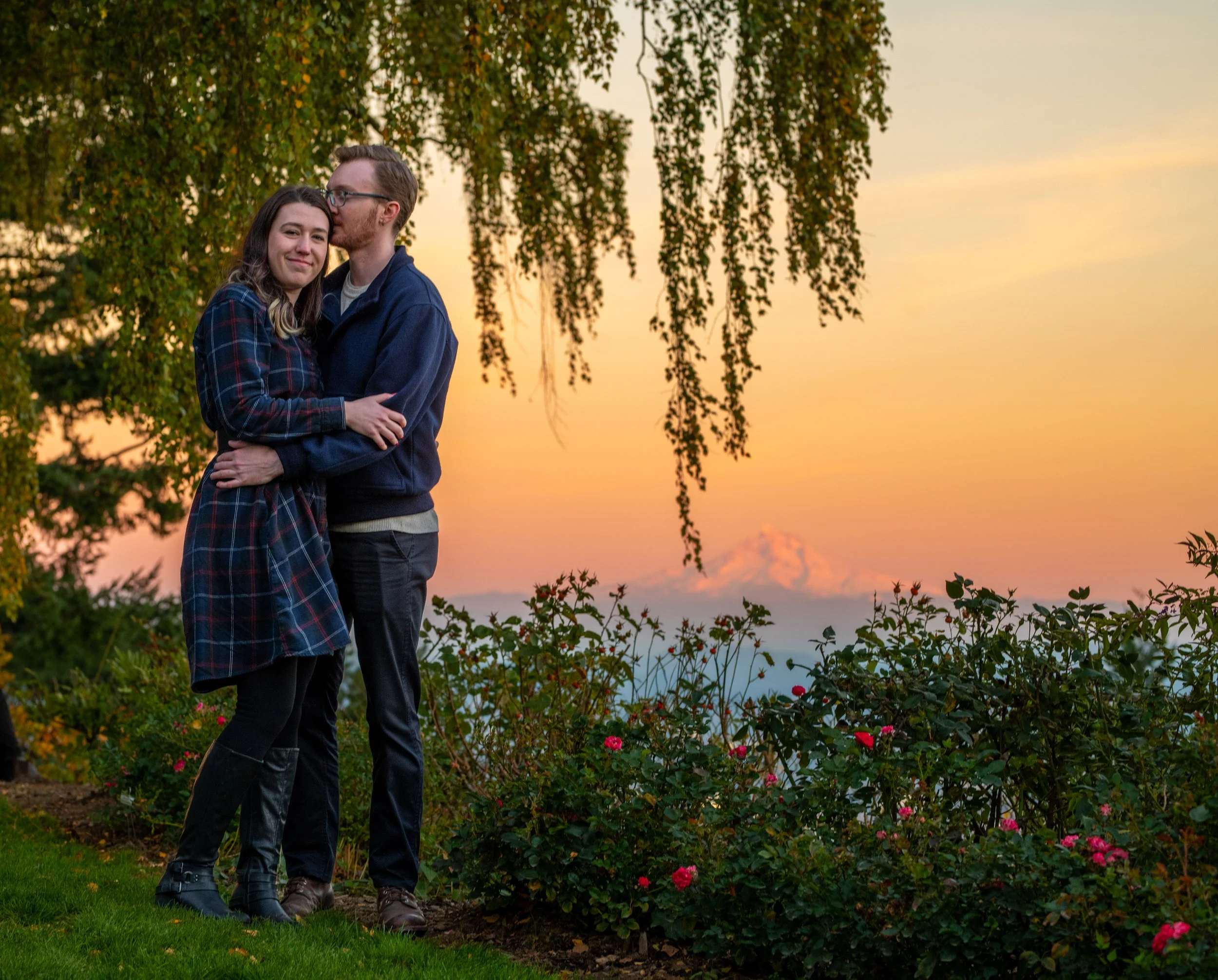 A couple hugging outdoors during sunset, with trees and flowering bushes in the background.