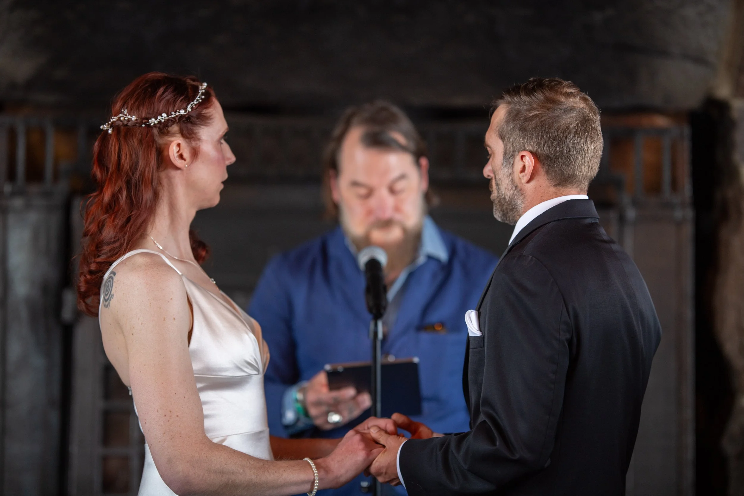 A woman with red hair and a tattoo on her shoulder, wearing a white dress and a pearl headband, is standing with a man with short gray hair and a beard, dressed in a black suit and white shirt. They are holding hands and facing each other during a we