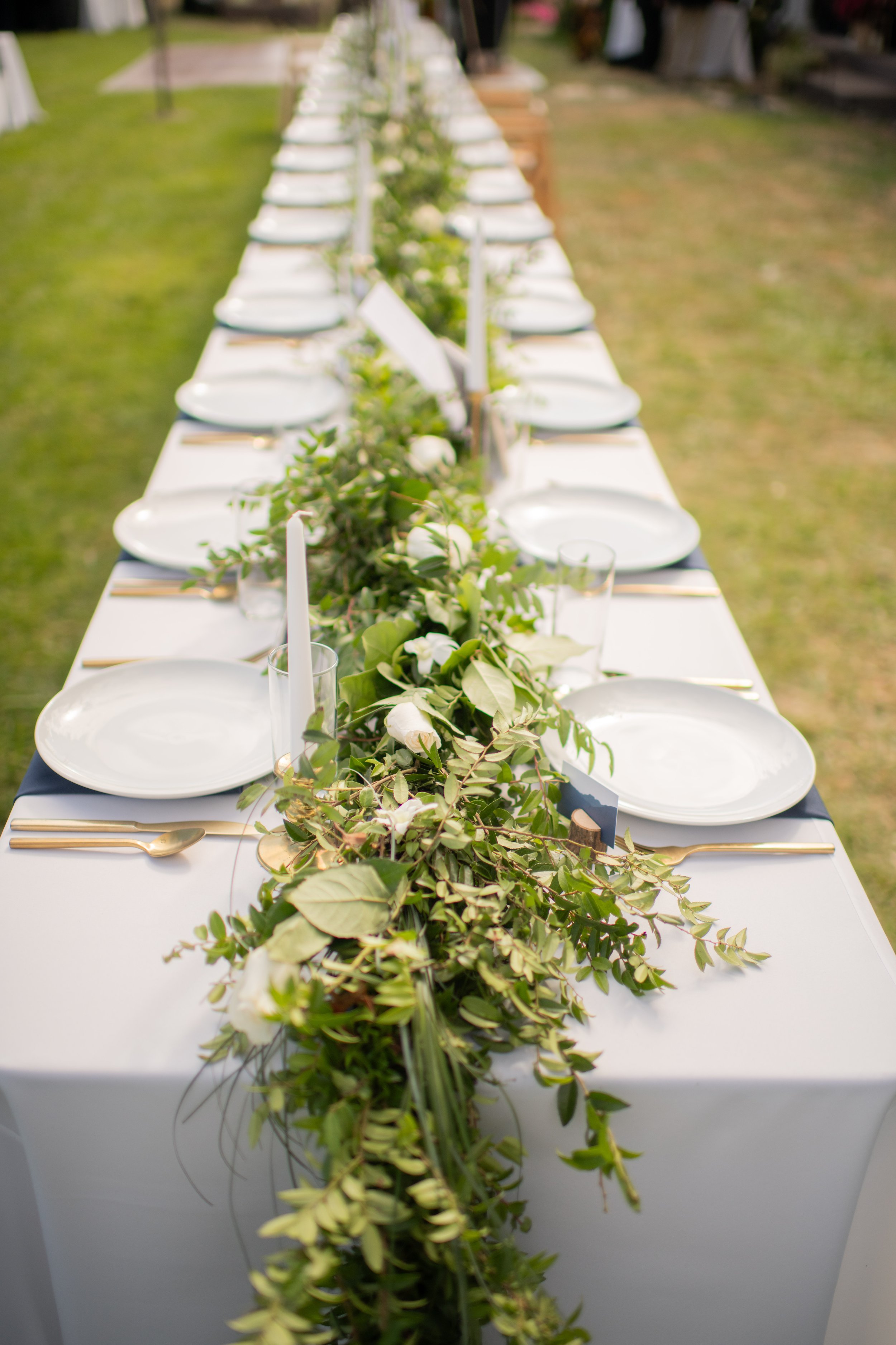 A long rectangular table set for an outdoor event, decorated with green foliage, white flowers, white plates, gold utensils, and glassware, with a green grassy area in the background.