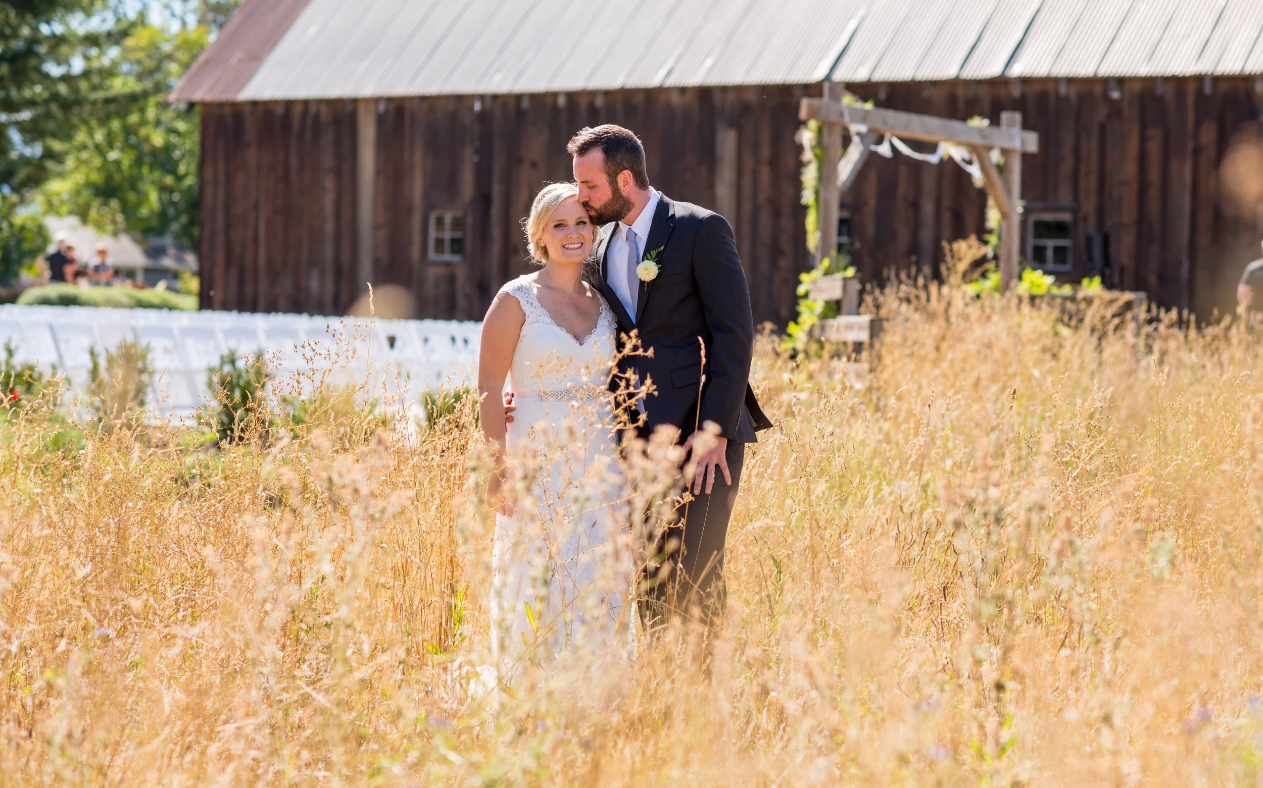 A bride and groom standing in a field of tall, dry grass during their outdoor wedding, with a rustic barn and a wooden wedding arch in the background, on a sunny day.