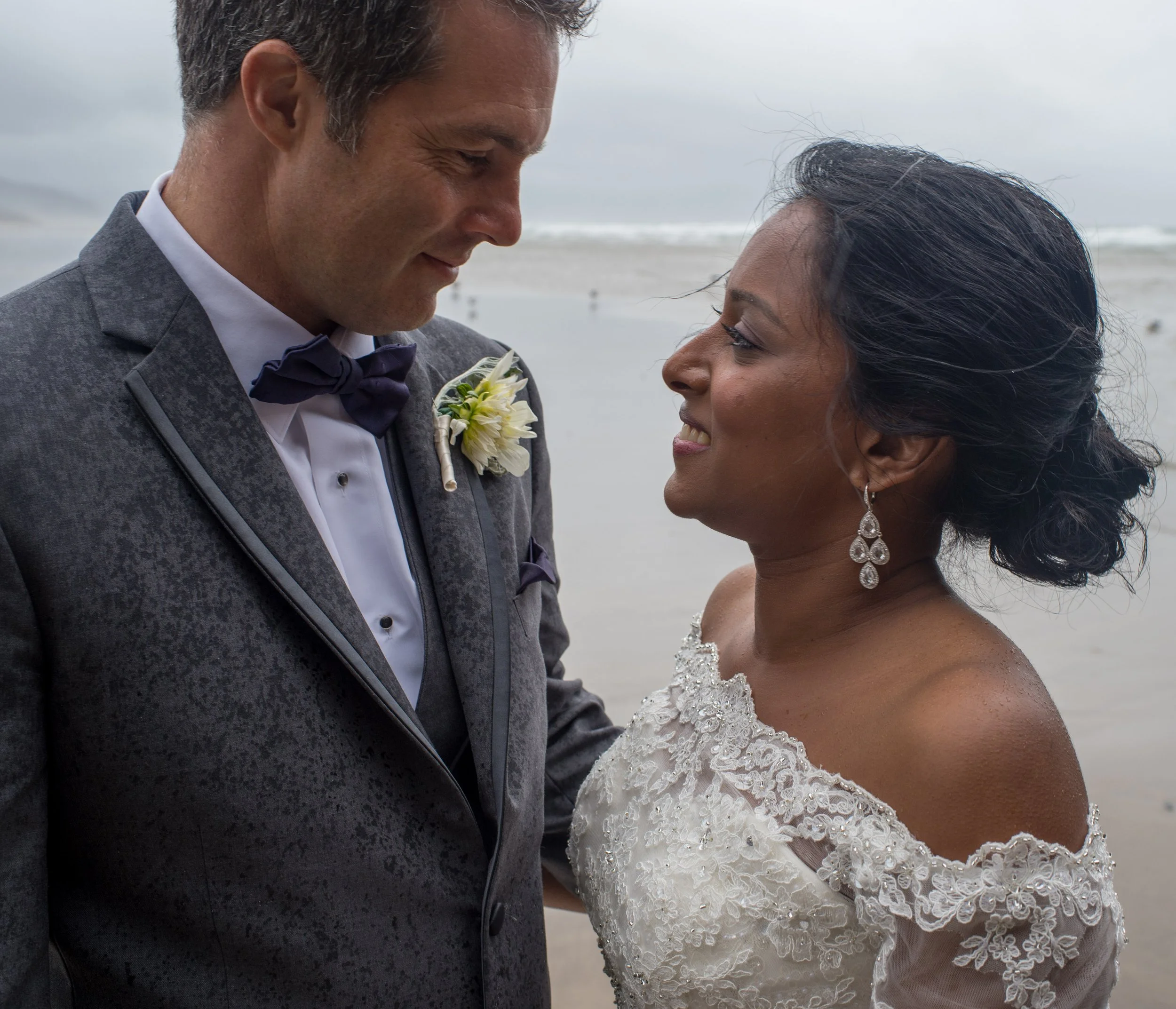 A bride and groom standing close on a beach, gazing at each other, with the groom wearing a grey suit and the bride in a lace wedding dress, holding hands.