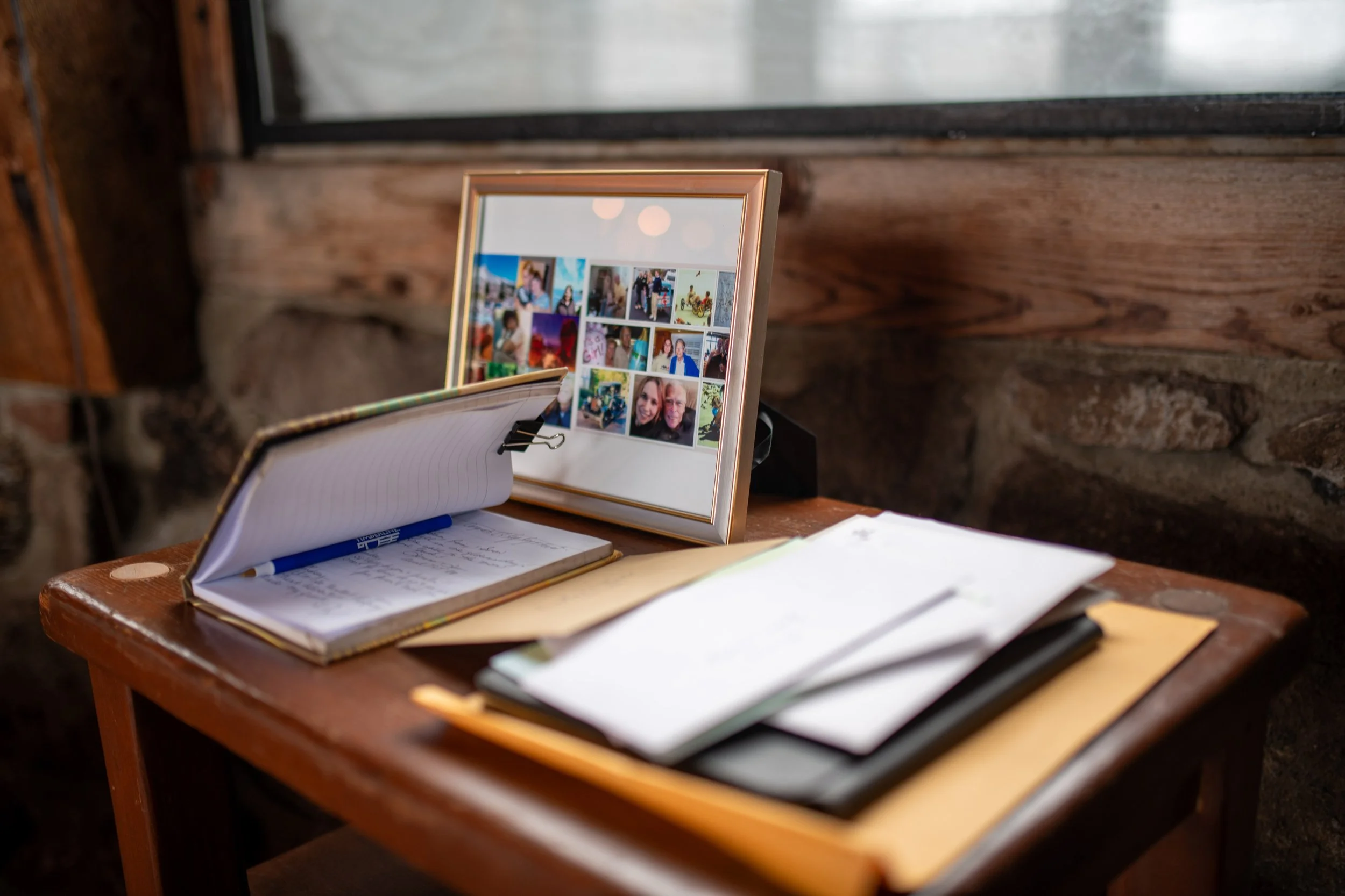 A small wooden table with an open notebook, blue pen, and a photo collage in a gold frame leaning against the wall. The photo collage contains numerous colorful photographs of people and moments.