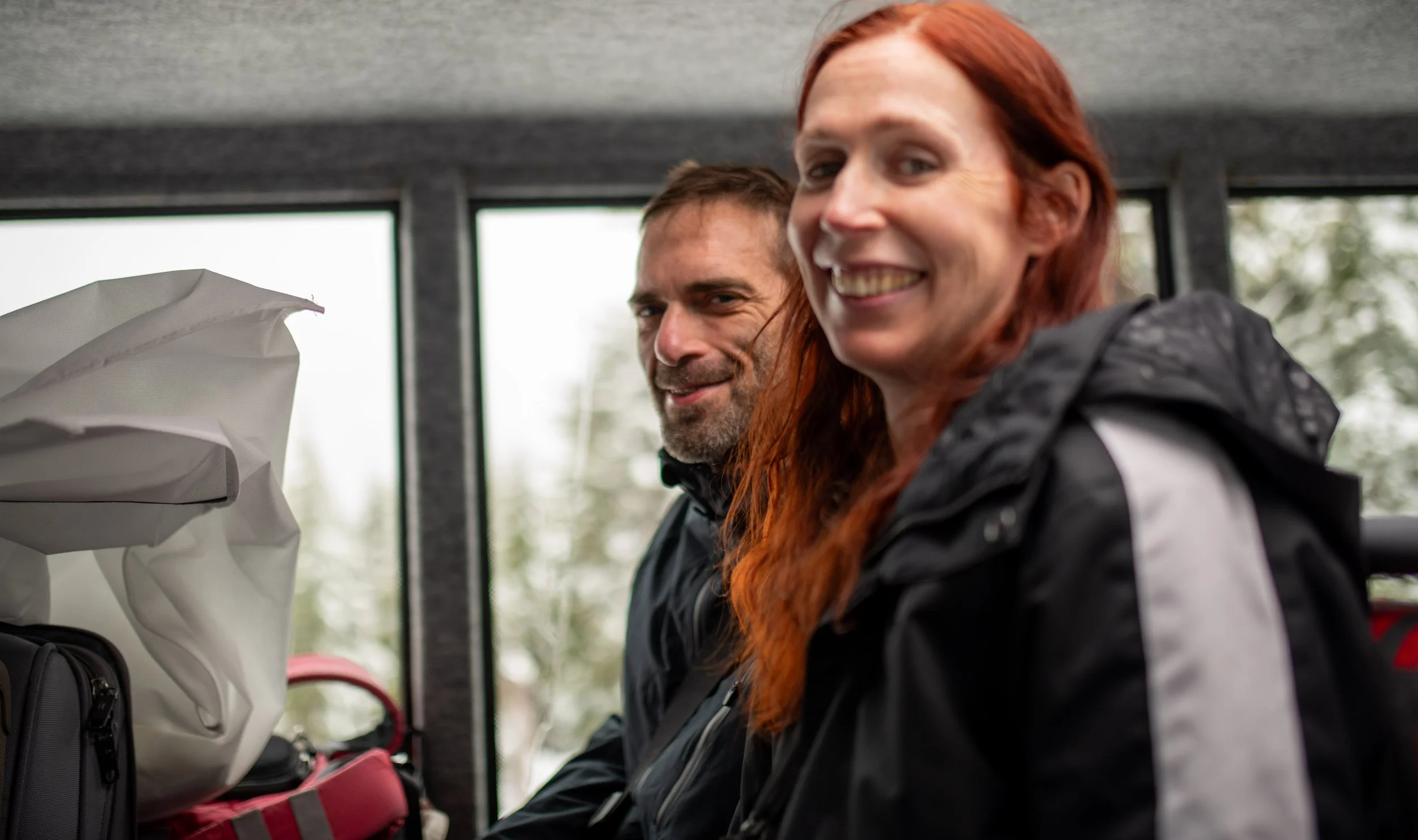 Two smiling people, a man and a woman, inside a golf cart with golf clubs and bags in the background.