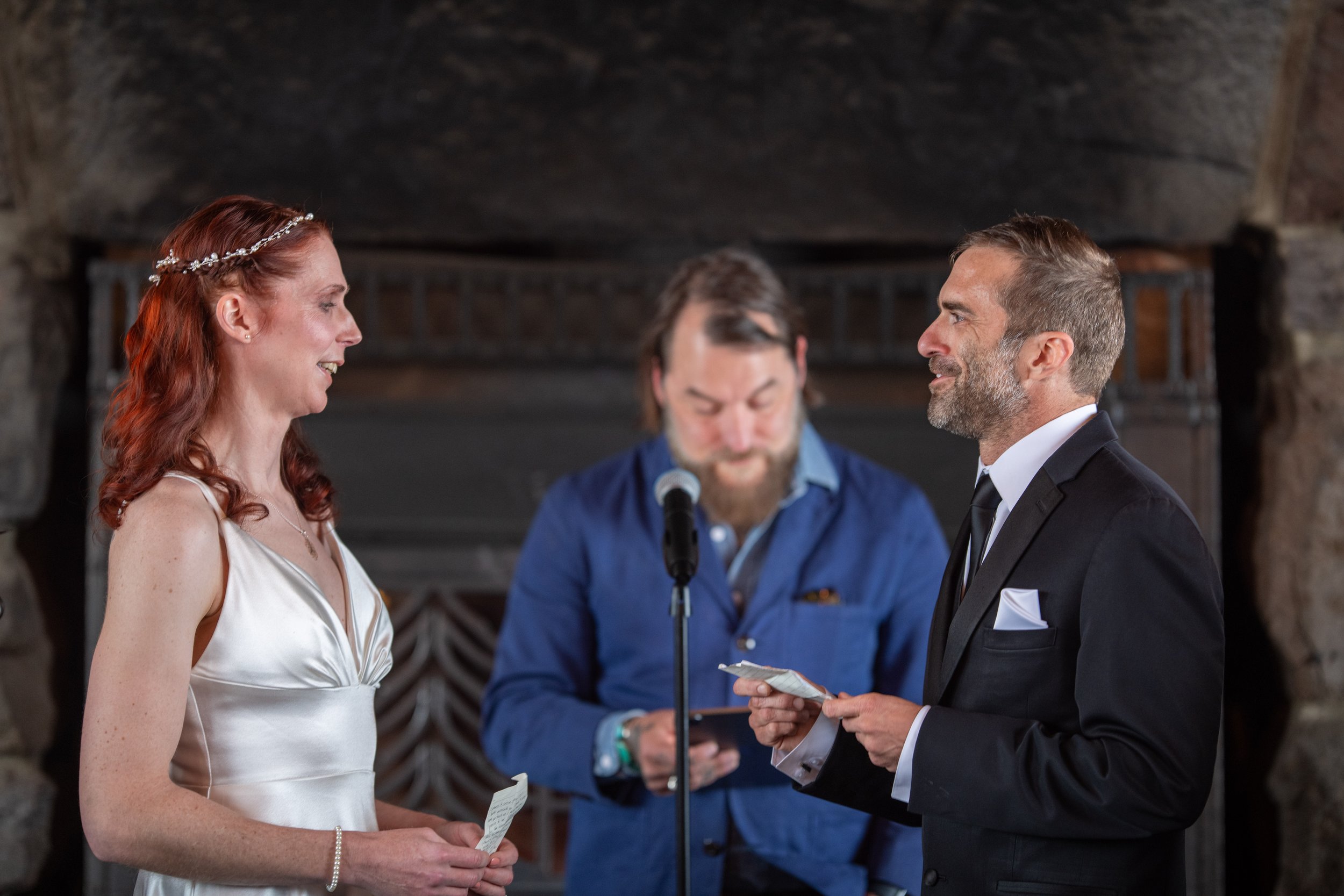 A groom and bride standing face-to-face during a wedding ceremony, with an officiant in the background reading from a paper, inside a rustic stone venue.