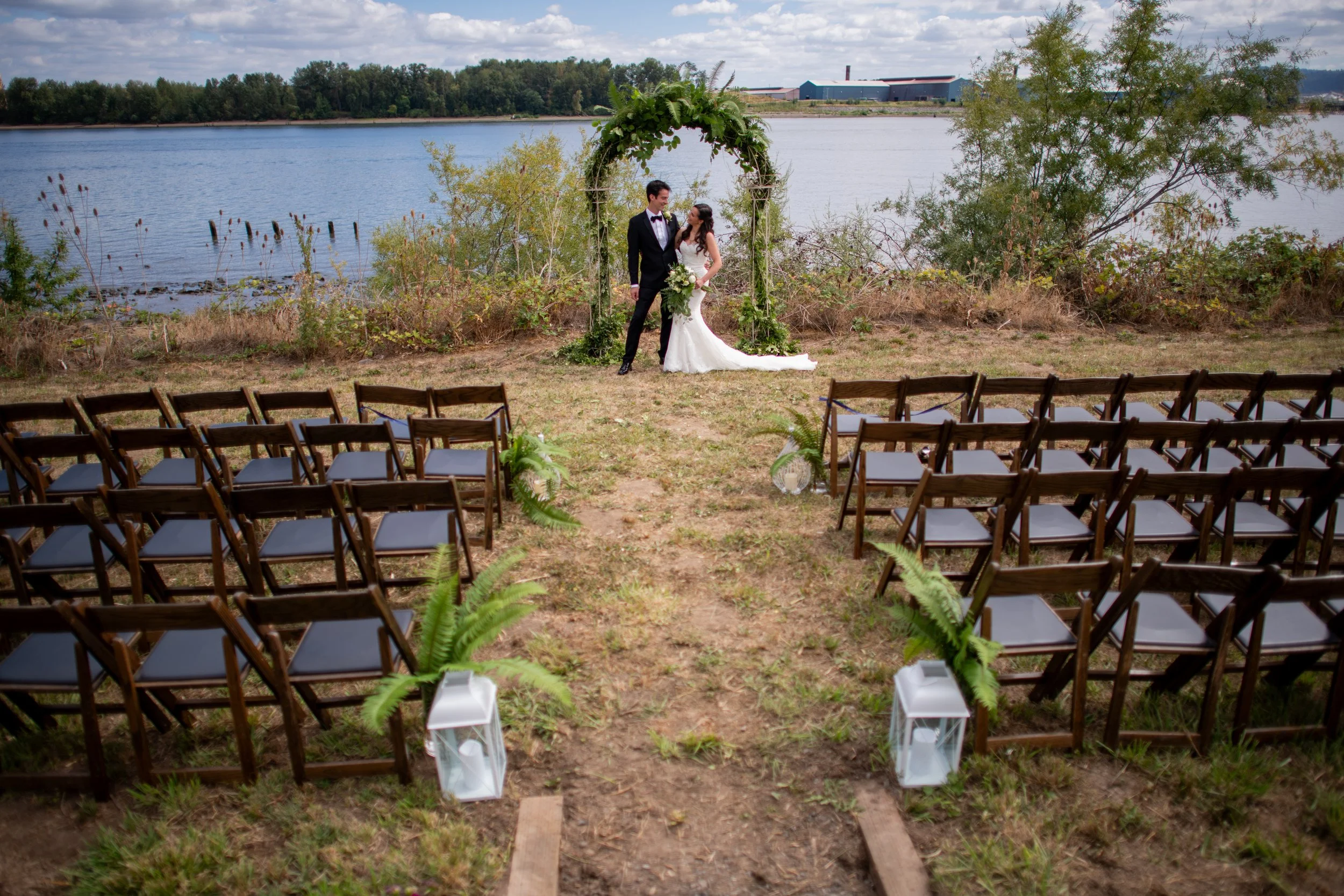 A bride and groom standing under a decorative arch by a body of water during their outdoor wedding ceremony, with chairs arranged for guests and trees in the background.