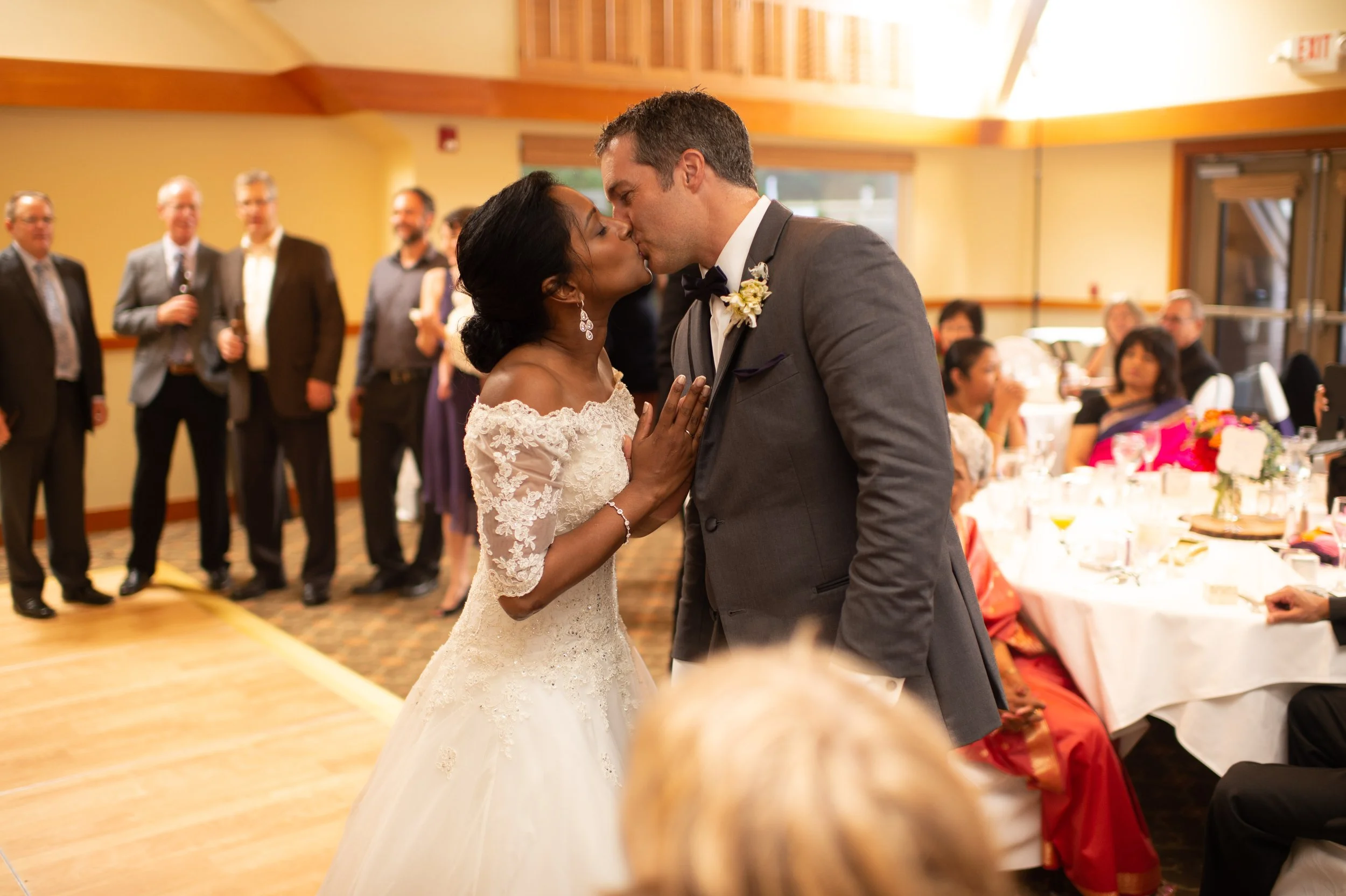 A bride and groom share a kiss during their wedding reception in a decorated event hall, with seated guests and friends in the background.