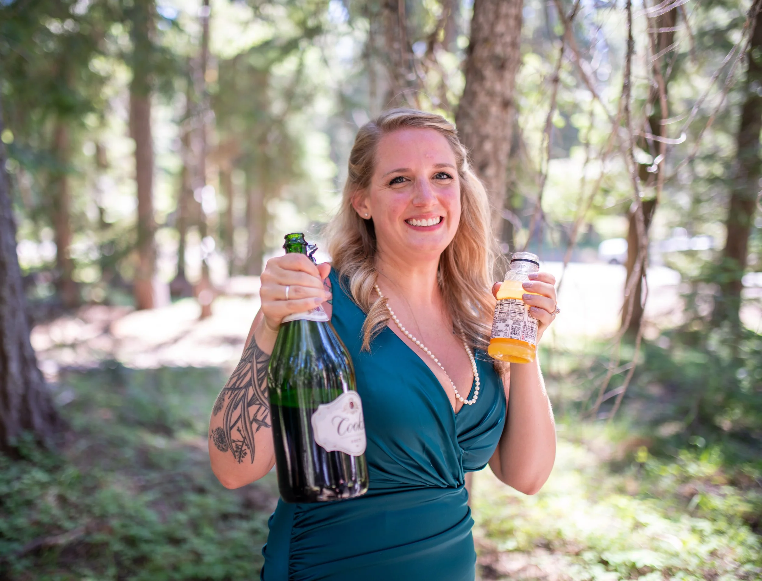 A woman with blonde hair smiling outdoors in a forest, holding a bottle of champagne and a bottle of orange juice.