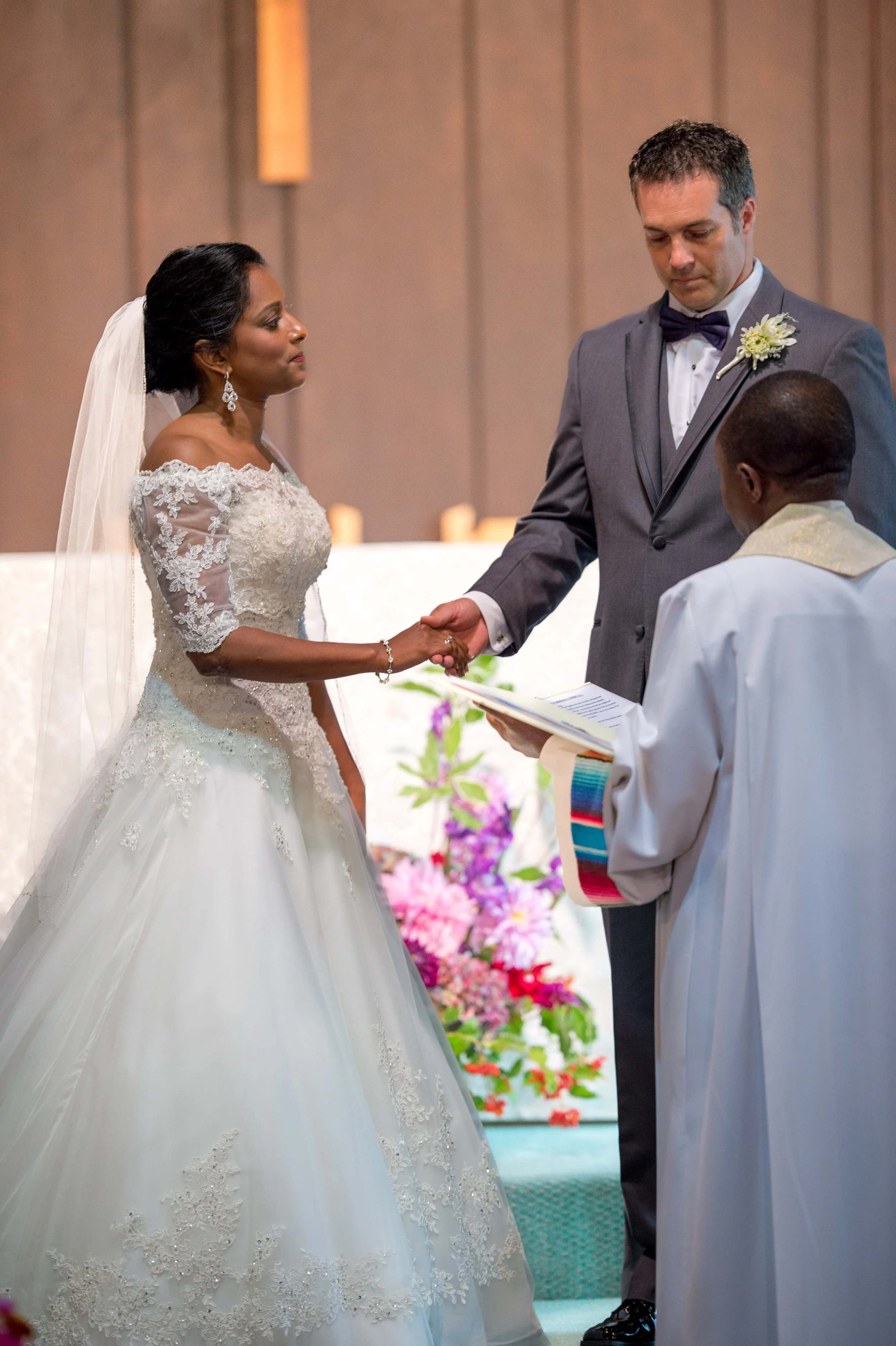 A wedding ceremony with an African American bride and a white groom. The bride is wearing a white lace wedding dress and the groom is in a gray suit with a purple bow tie. They are holding hands and facing each other, with an officiant standing in fr