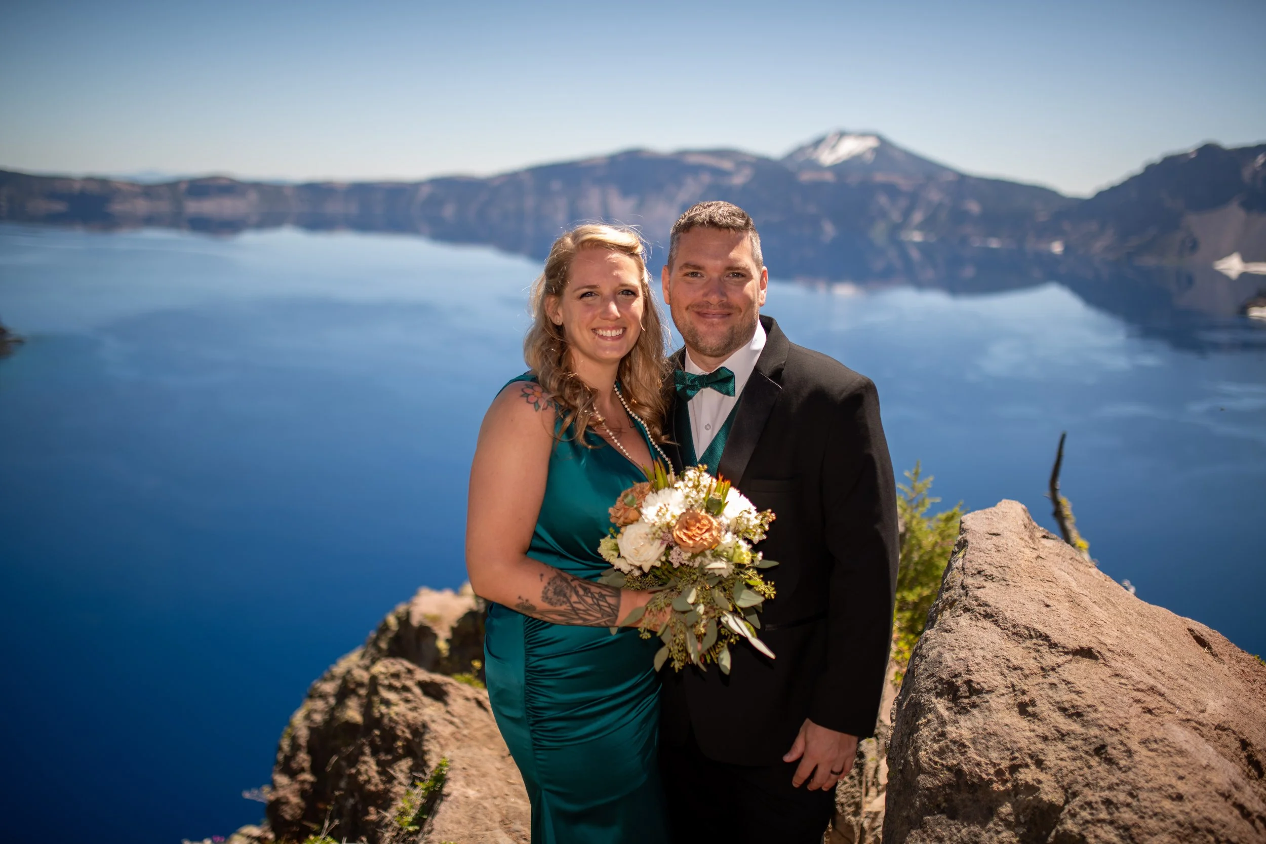 A smiling couple in formal attire, the woman holding a bouquet of flowers, standing on rocky terrain by a large lake with mountains in the background on a clear, sunny day.