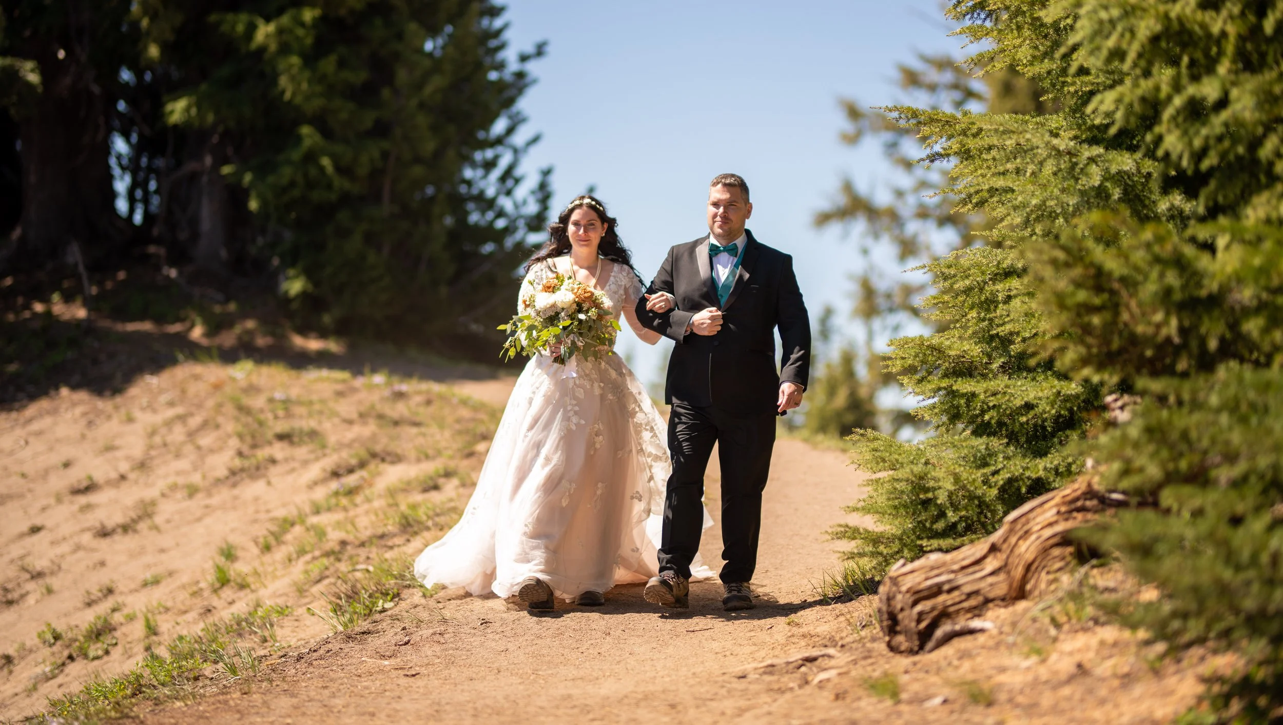 A bride and groom walking hand in hand outdoors on a dirt path with trees on both sides, the bride holding a bouquet of flowers and wearing a lace wedding dress, and the groom dressed in a black tuxedo with a teal bow tie, under clear blue sky.