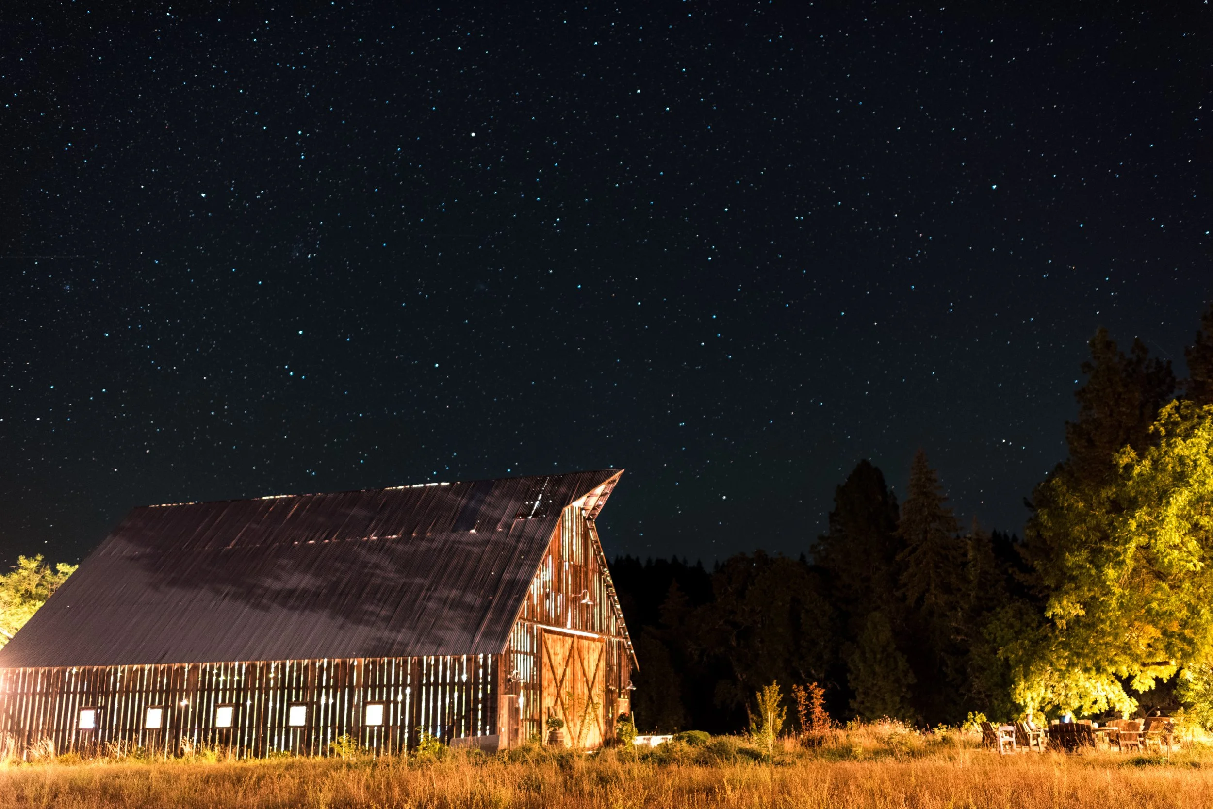 Nighttime scene with a star-filled sky and a rustic barn with lights. Trees are visible in the background.