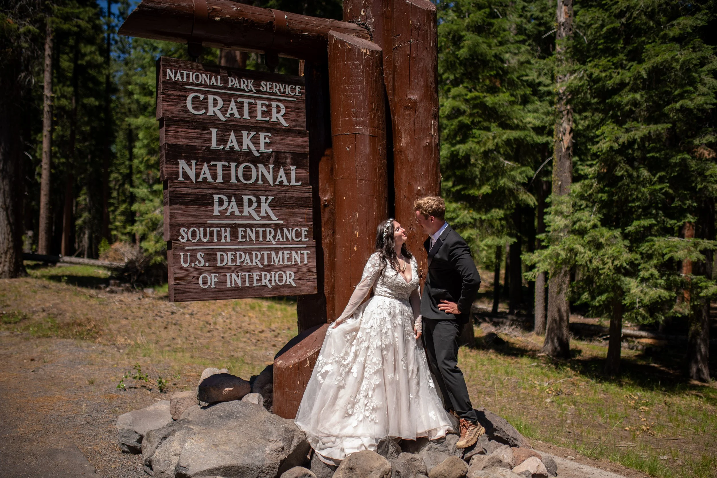 A bride and groom in wedding attire standing close together in front of a large wooden sign for Crater Lake National Park, surrounded by tall green trees.