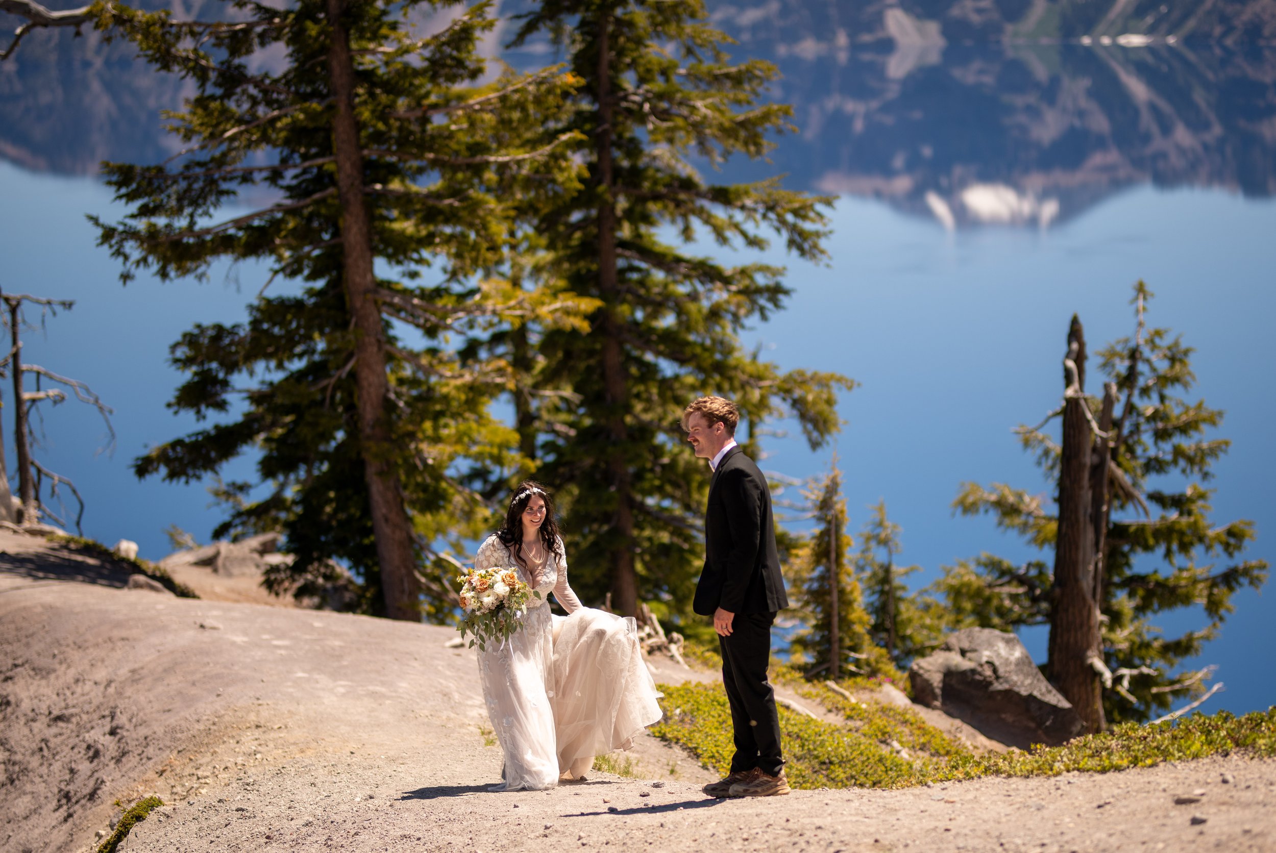 A bride and groom standing outdoors on a mountainous lakeside landscape during their wedding, with tall trees and a calm lake in the background.