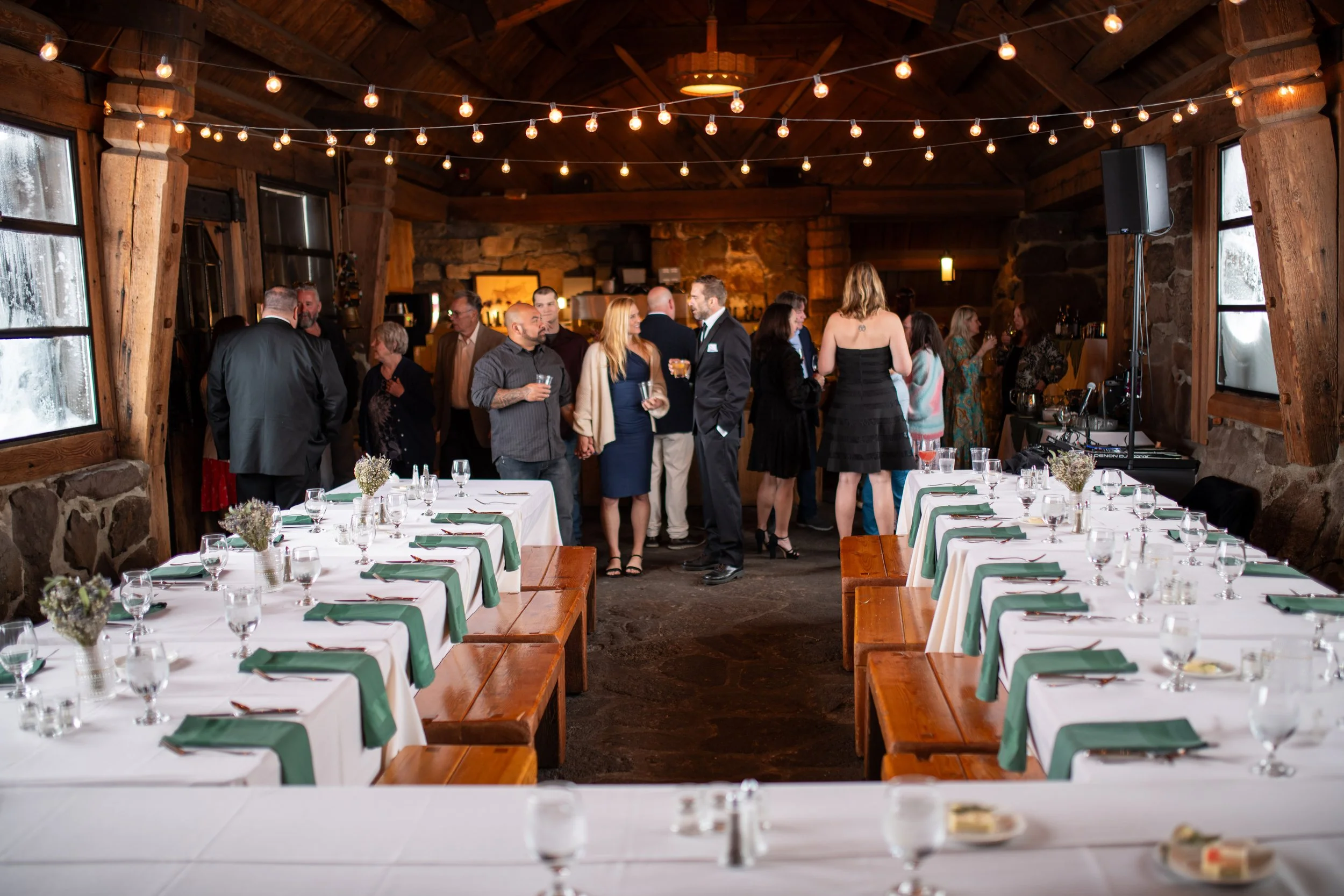 People socializing at a rustic indoor event with wooden walls and string lights, with tables set with plates and glasses.