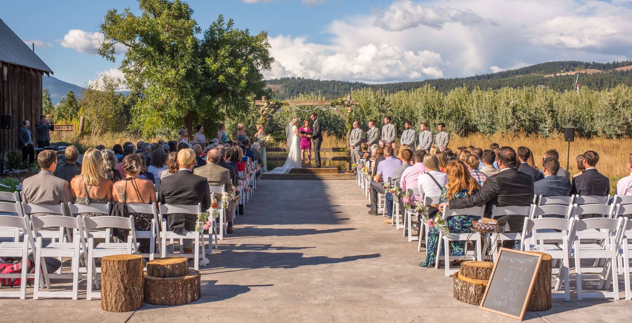 An outdoor wedding ceremony taking place on a sunny day with a scenic mountain and forest backdrop. The bride and groom stand under a rustic wooden arch decorated with flowers, exchanging vows. Guests sit in white chairs decorated with pink flowers, 
