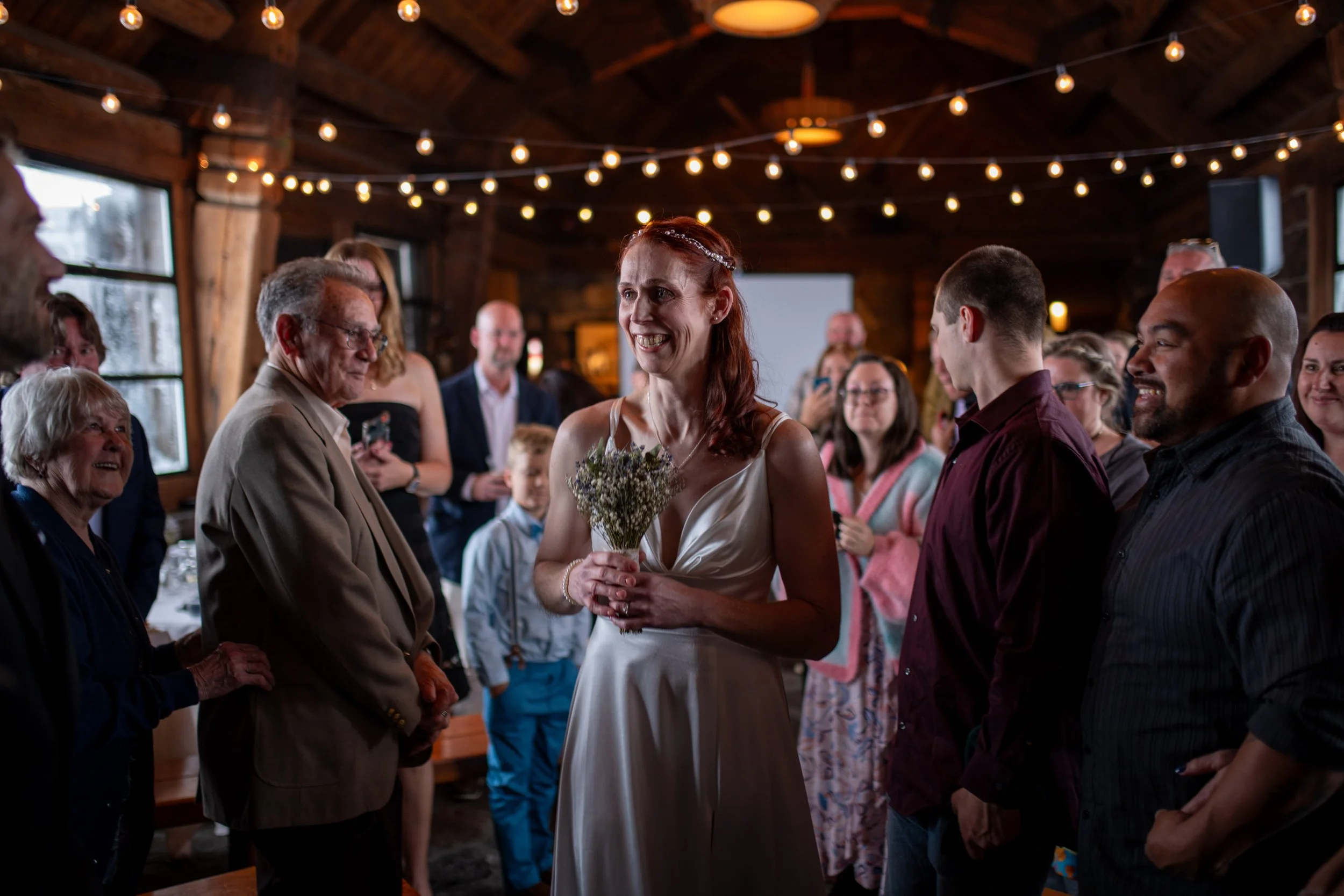 A group of people, including a woman in a white dress holding a small bouquet, gathered indoors with string lights overhead, celebrating a wedding or special occasion.