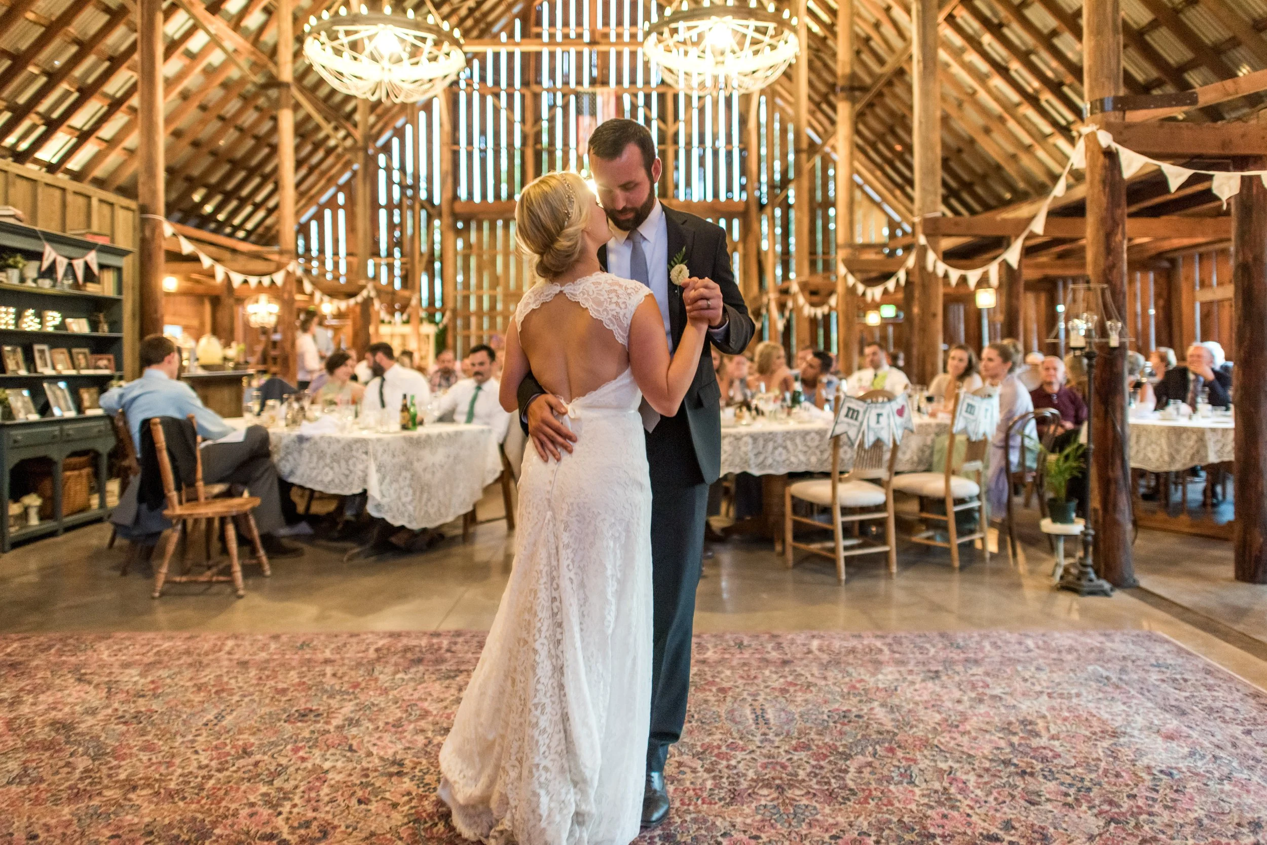 A bride and groom dancing at their wedding in a rustic barn with wooden beams, chandeliers, and guests seated at decorated tables in the background.