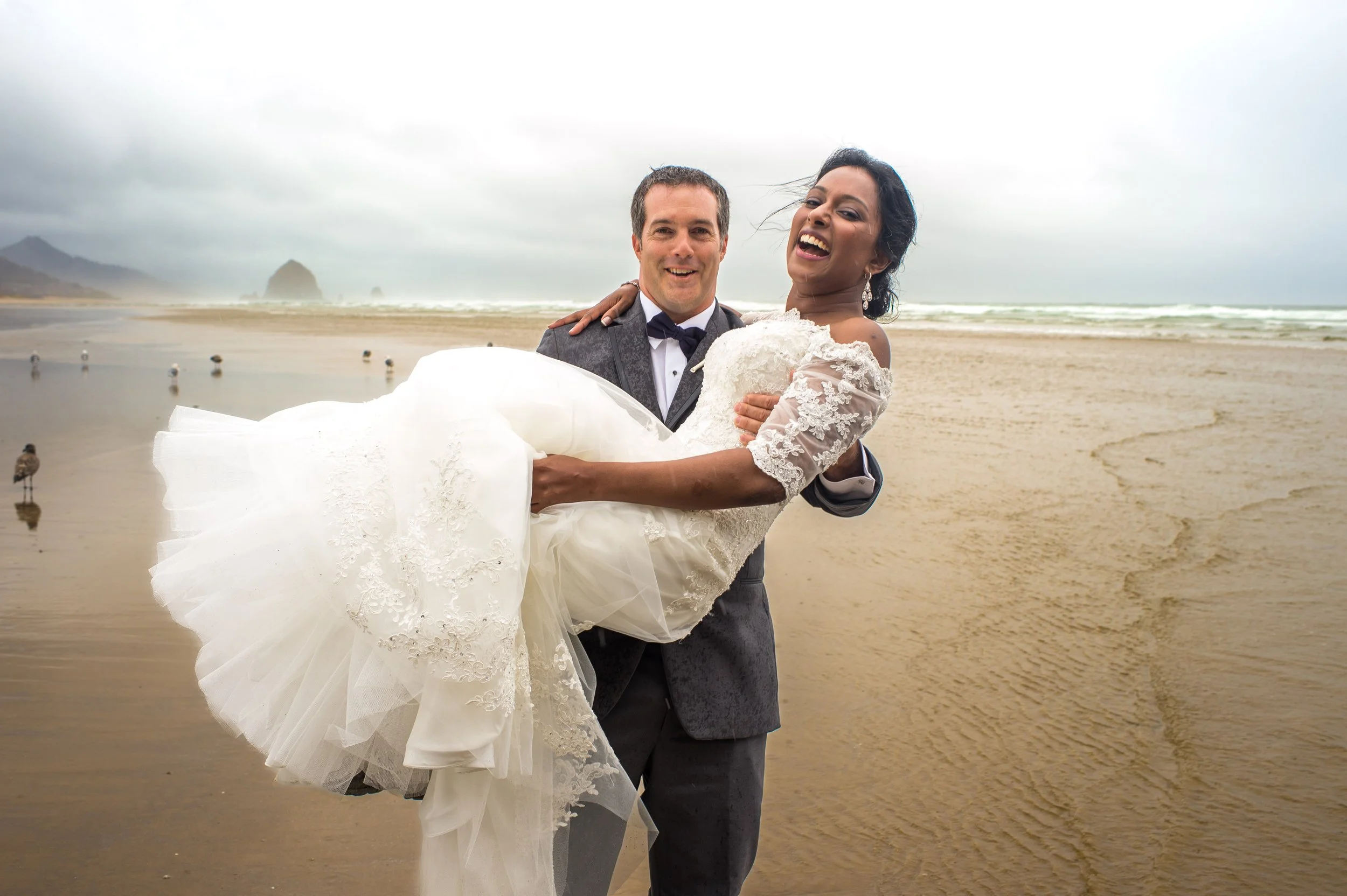 A groom holding a bride in a white wedding dress on a beach with cloudy sky and distant cliffs.