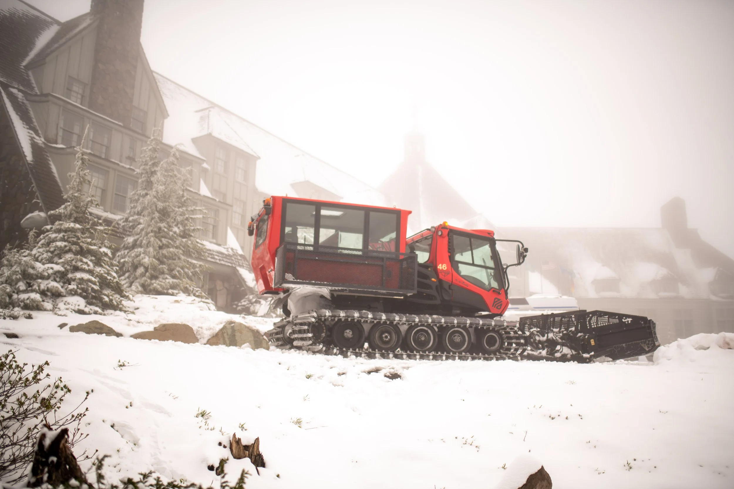 A red snow grooming machine on a snowy slope in front of a large house with snow-covered trees and foggy weather.