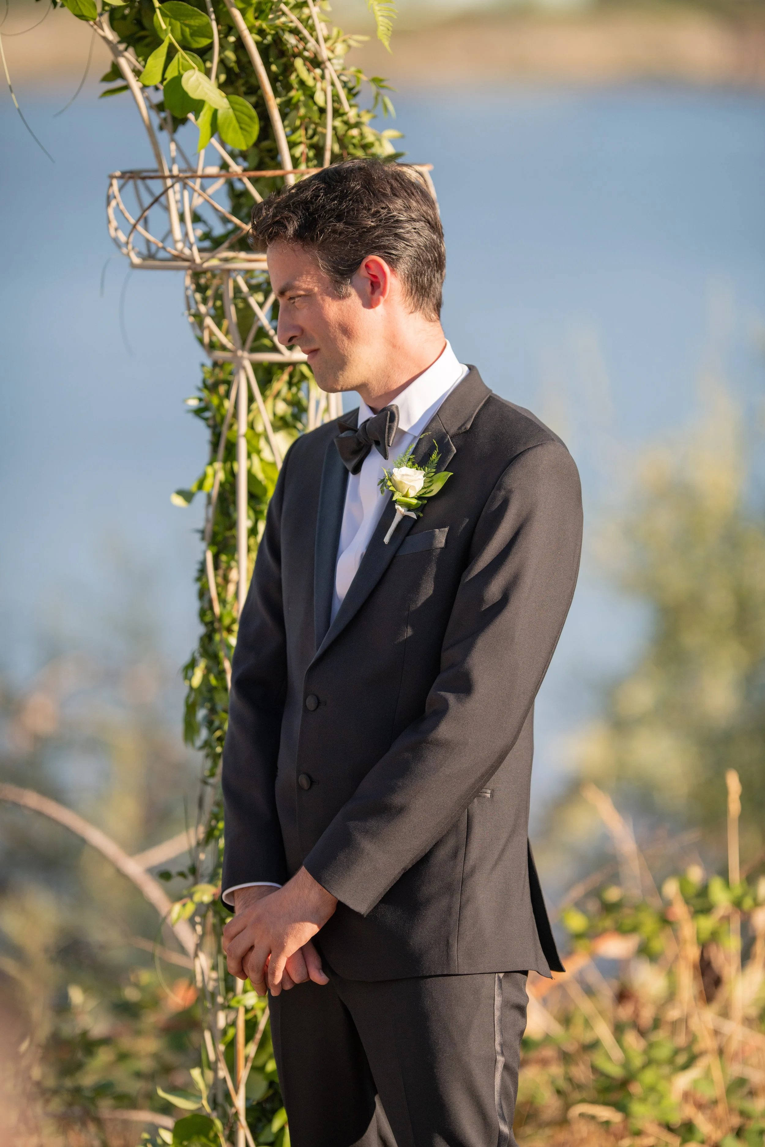 A man in a black tuxedo with a bow tie and a white boutonniere on his lapel standing outdoors near a body of water.