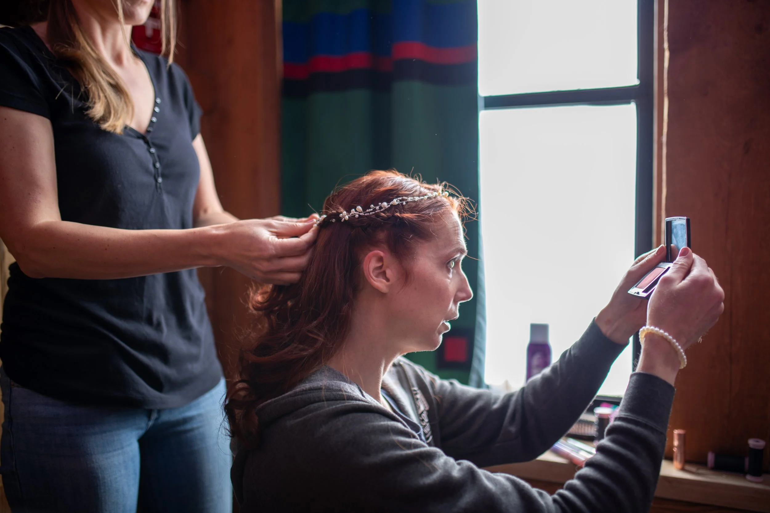 A woman with red hair styled with a pearl headband sitting at a makeup station taking a selfie with her phone, while another woman, dressed in dark clothing, adjusts her hair. The setting appears to be indoors with natural light coming through a wind
