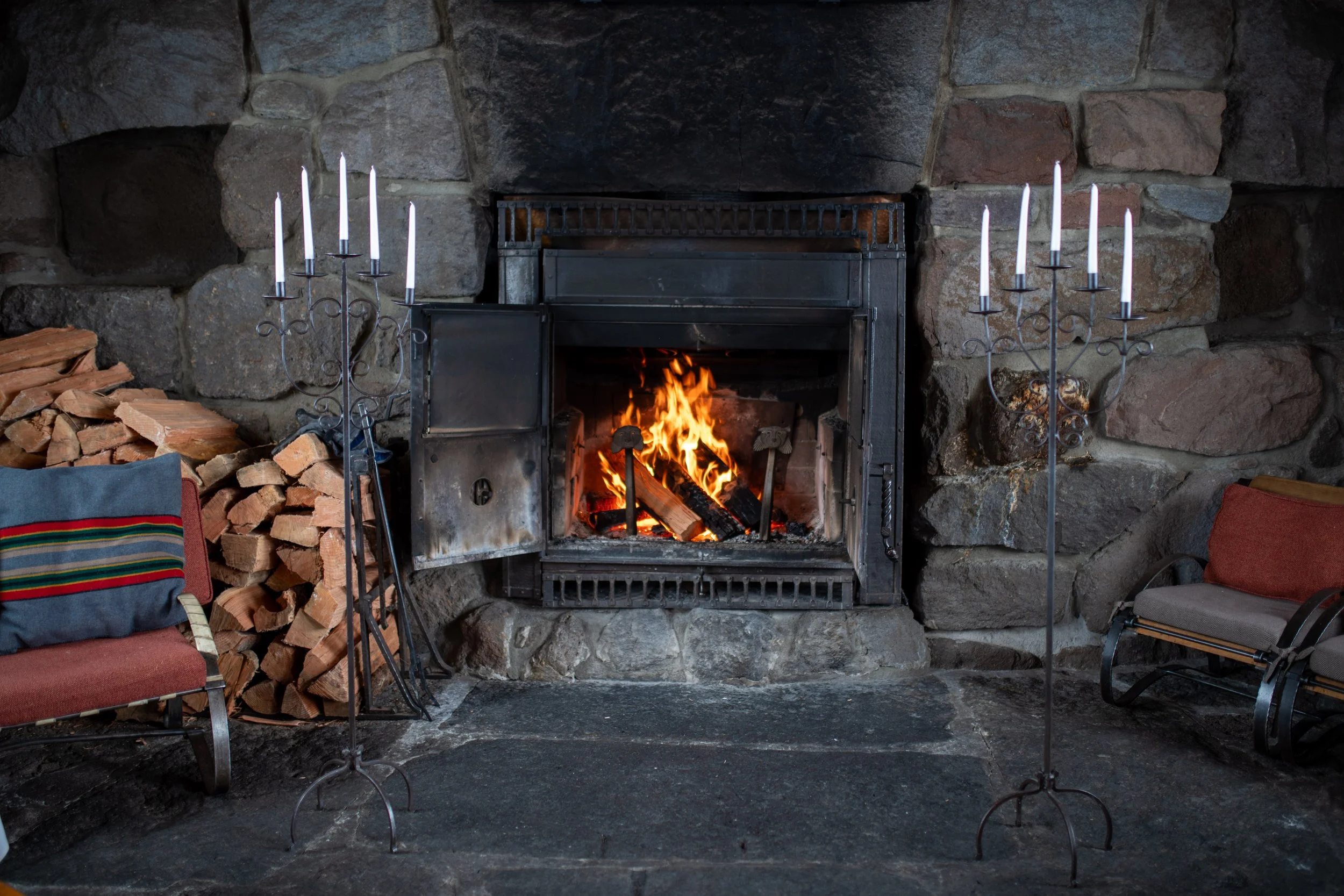 A cozy wood-burning fireplace with a fire burning, surrounded by a stone wall. Two candelabra with white candles are positioned on each side of the fireplace, and there is a stack of firewood on the left. Two chairs are partially visible, one on each