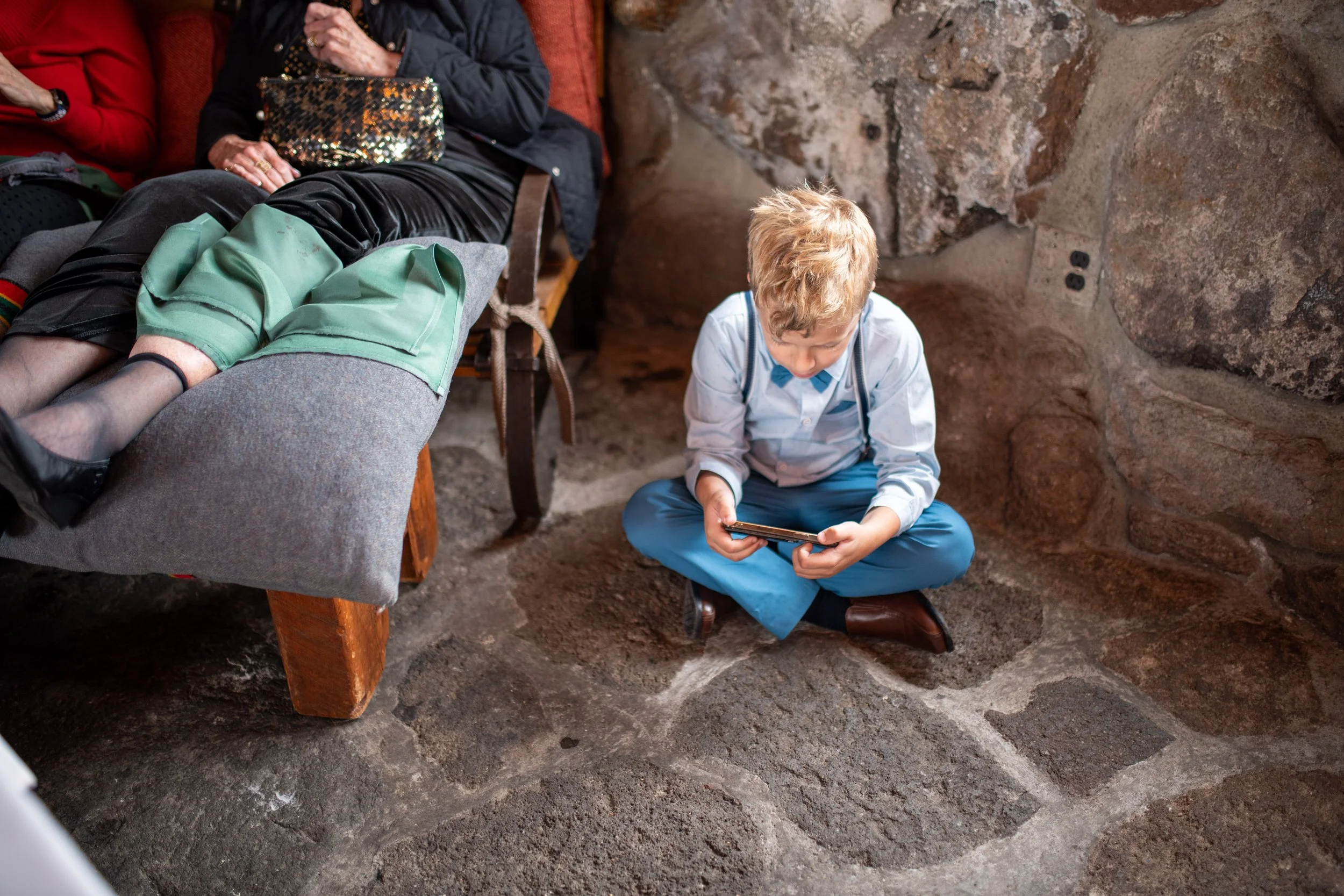 A young boy with blond hair sitting on the ground inside a cave, looking at a smartphone. An adult is lying on a cushioned bench, with legs crossed, partially visible on the left, with colorful socks, black skirt, and a dark jacket. The cave has rugg