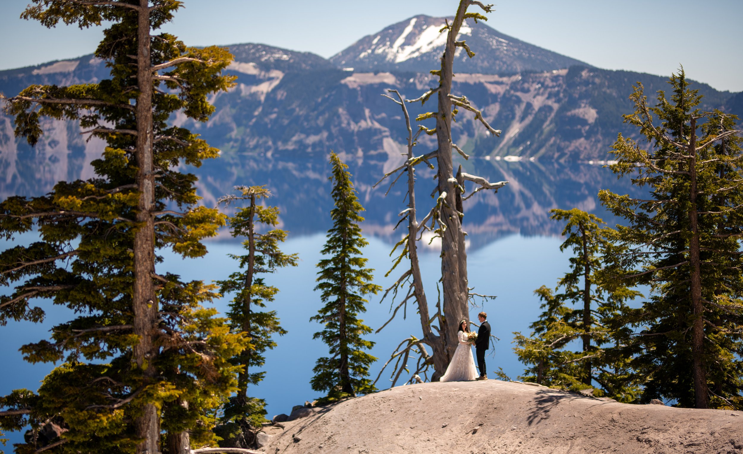 A bride and groom stand together on a small hilltop surrounded by tall trees, with a mountain with snow patches in the background and a lake reflecting the mountain.