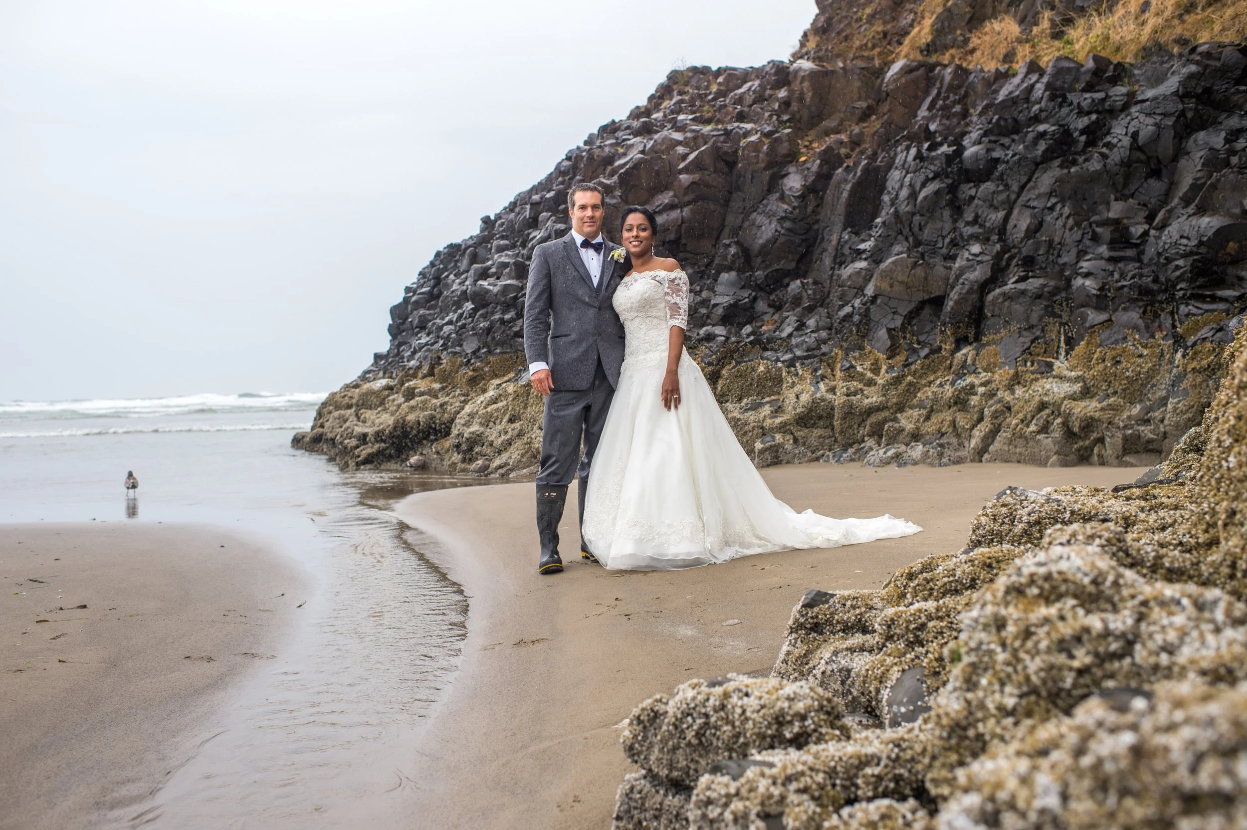 A wedding couple in formal attire standing on a beach near rocky formations, with the ocean and a cloudy sky in the background.