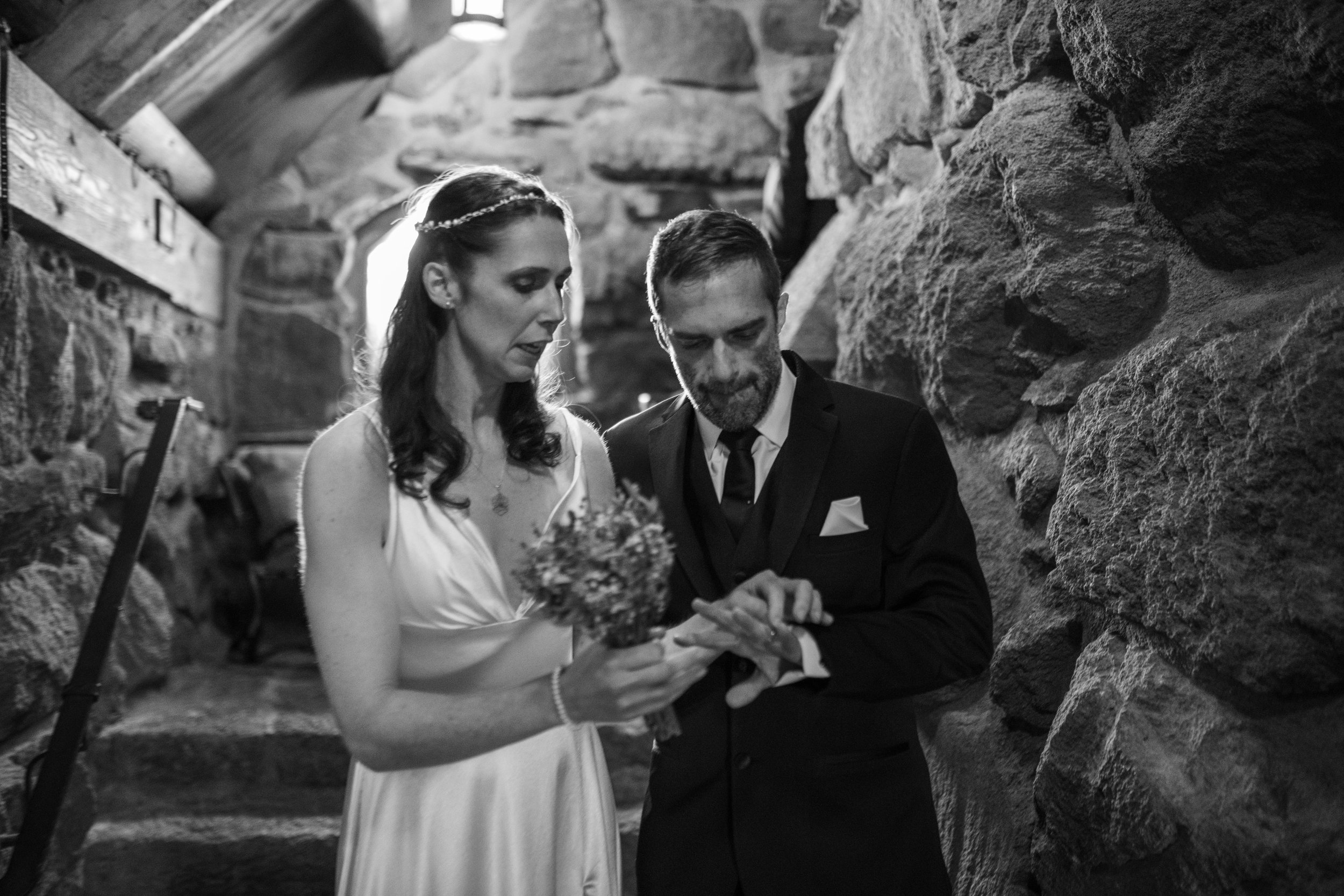 A black and white photo of a bride and groom in wedding attire, looking at something together and smiling, against a stone wall background.