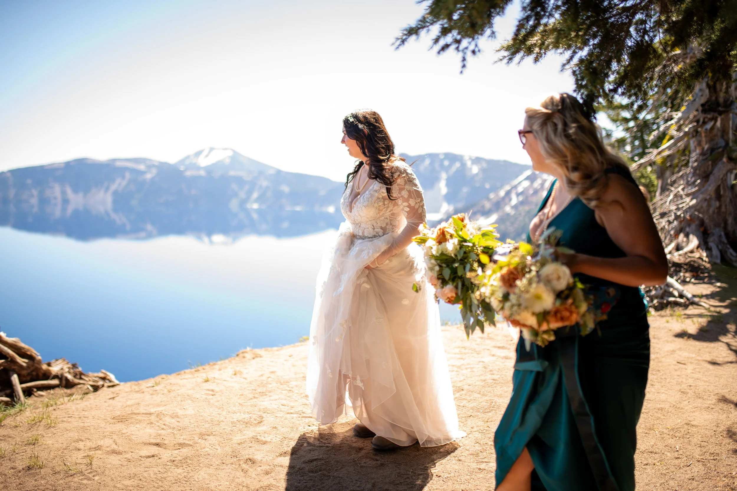 A bride in a wedding dress standing outdoors by a lake with snow-capped mountains in the background, talking to a woman holding a bouquet of flowers, under a tree with the sun shining.