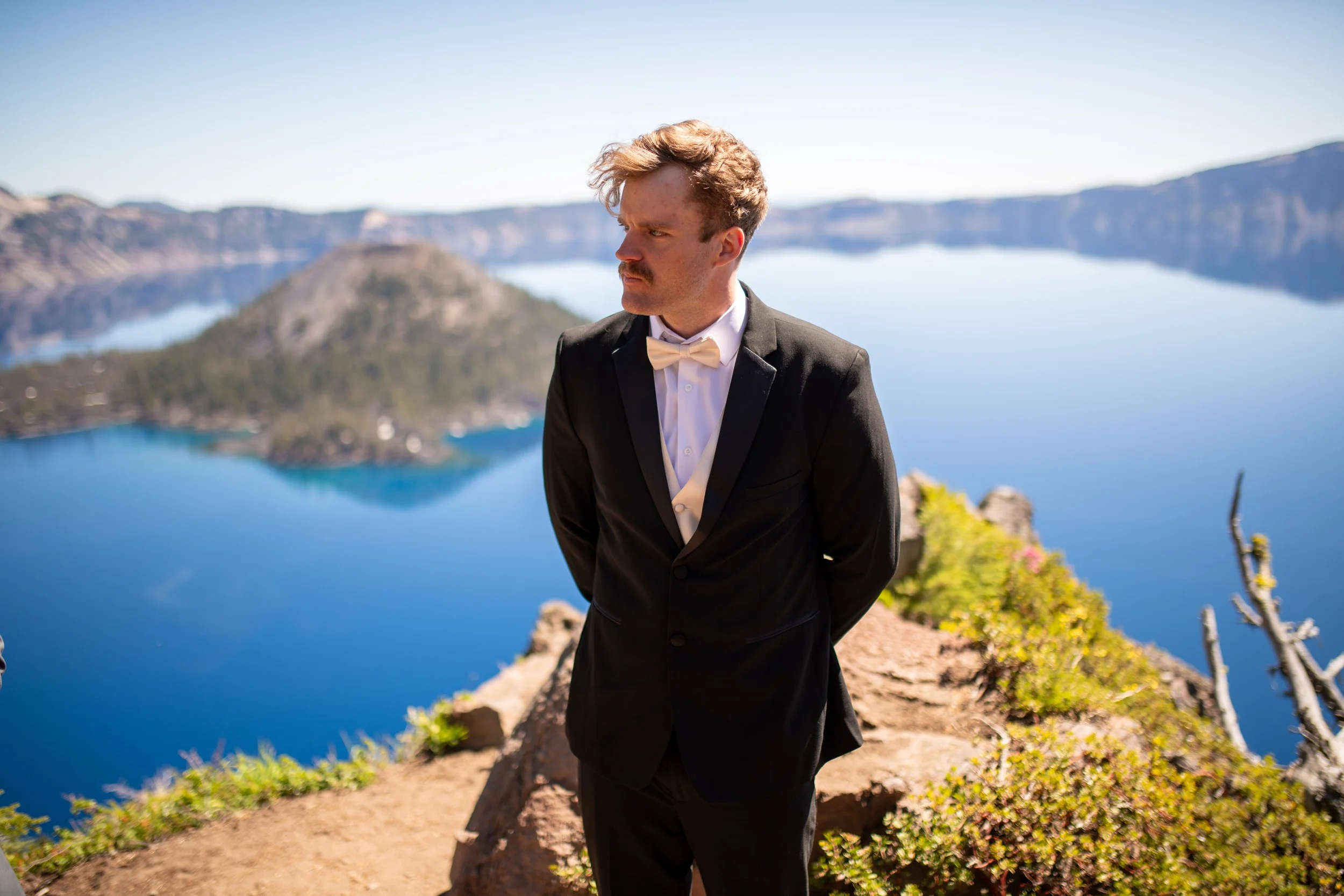 A man dressed in a tuxedo with a bow tie stands outdoors near a lake with canyon walls in the background.